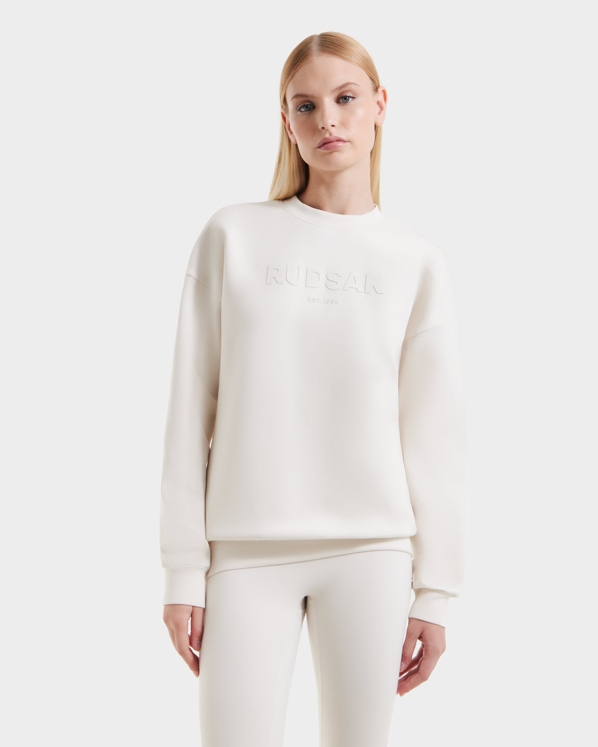 A person with long blonde hair models the Rudsak BRYN - 9524007 Vanilla W cotton crewneck sweatshirt, featuring embossed branding, styled with matching white pants against a plain white background.