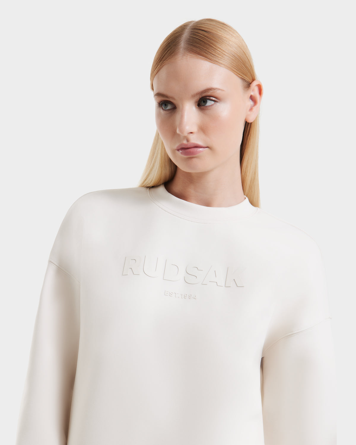 A woman with straight blonde hair and blue eyes wears the Rudsak BRYN - 9524007 Vanilla W, a white genderless cotton crewneck sweatshirt embossed with RUDSAK EST. 1994, standing against a plain white background.