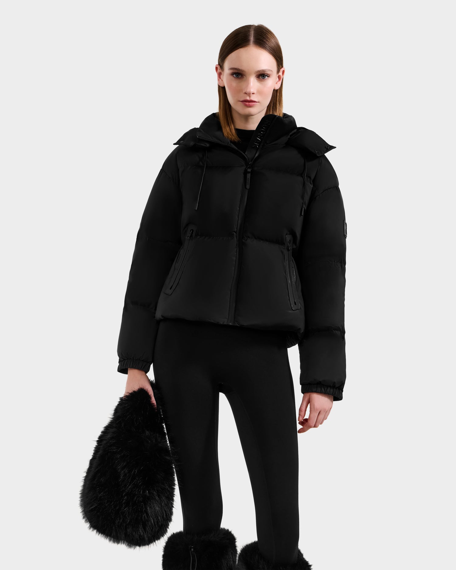 A woman with straight hair and a neutral expression stands against a plain background, dressed in winter layers and holding the Rudsak CABAS - 8325545 Black faux fur mini tote—a chic statement accessory.