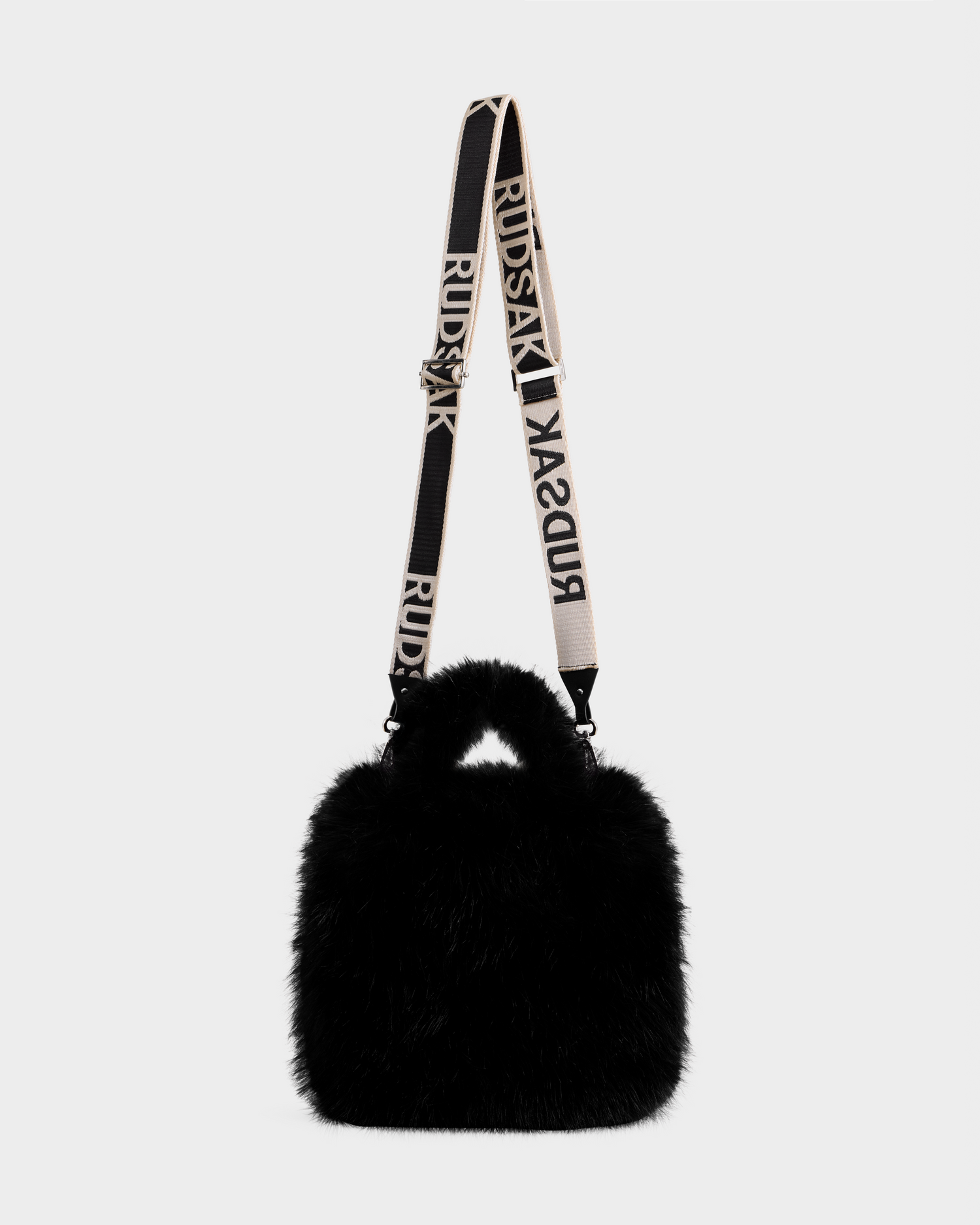 The Rudsak CABAS - 8325545 Black is a black faux fur mini tote with a short handle and a detachable beige RUDSAK-branded strap, set on a light gray background—an ideal winter statement accessory.
