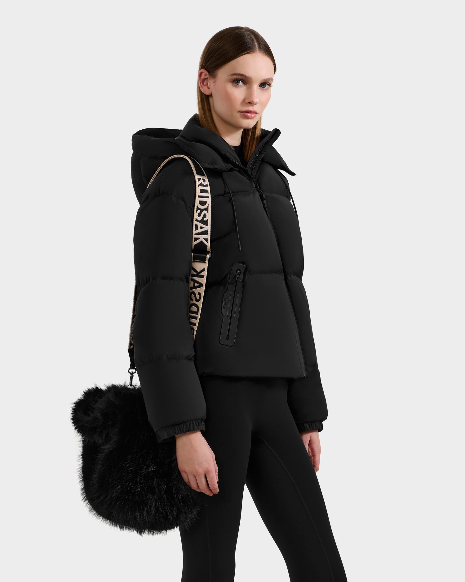 A woman in winter layers—black puffer jacket and leggings—poses against a plain background, carrying the Rudsak CABAS - 8325545 Black mini tote with a beige strap as a statement accessory over her shoulder.