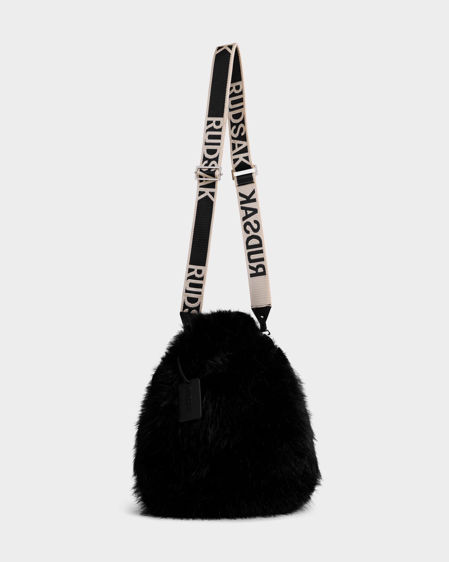 The CABAS - 8325545 Black by Rudsak is a black faux fur mini tote crossbody with a wide, adjustable strap stamped with “RUDSAK” in bold—an eye-catching accessory shown against a simple light backdrop.
