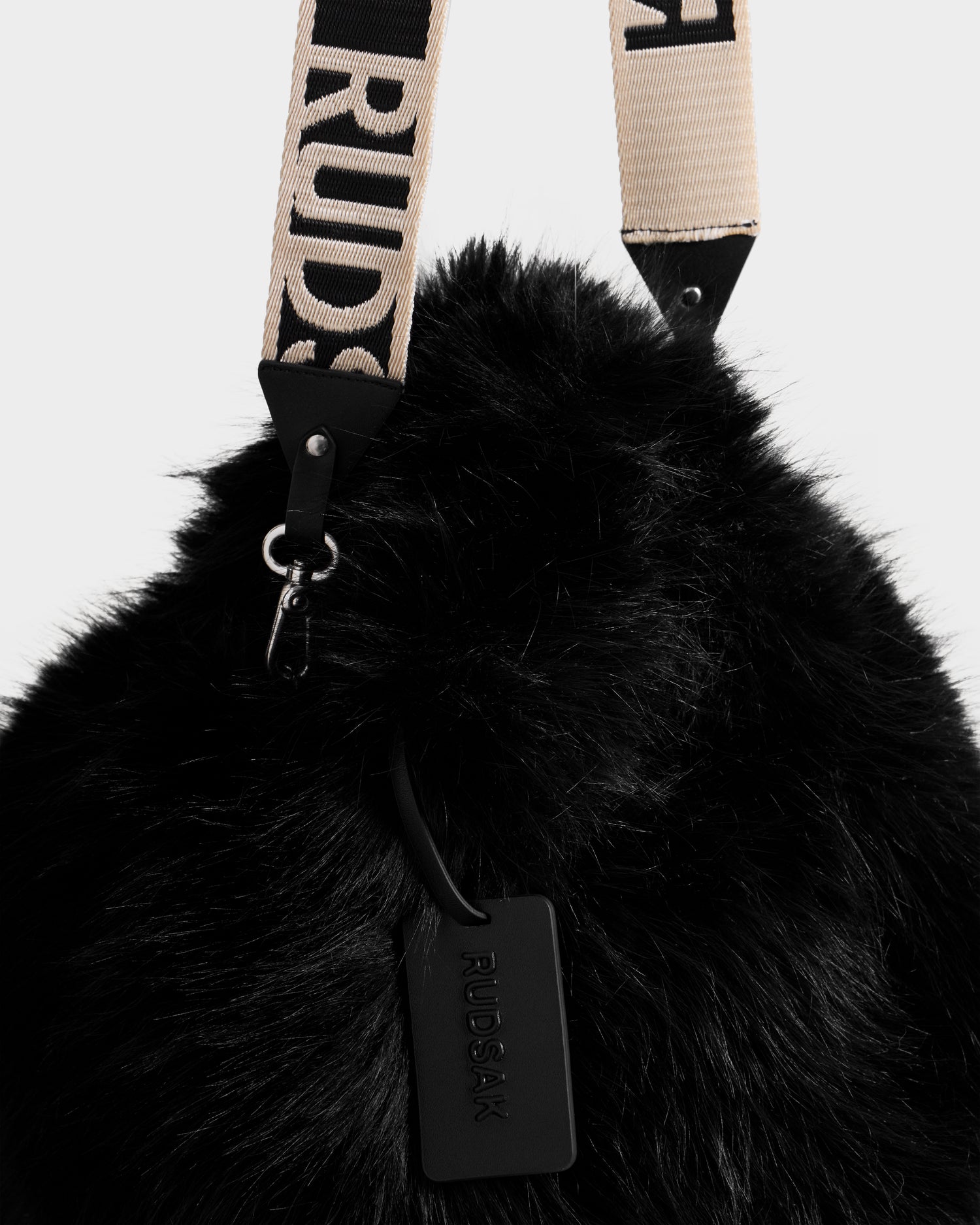 The CABAS - 8325545 Black by Rudsak is a black faux fur mini tote with a beige strap featuring “RUDSAK” in bold. A black leather Rudsak tag completes this statement accessory, ideal for elevating your winter look.