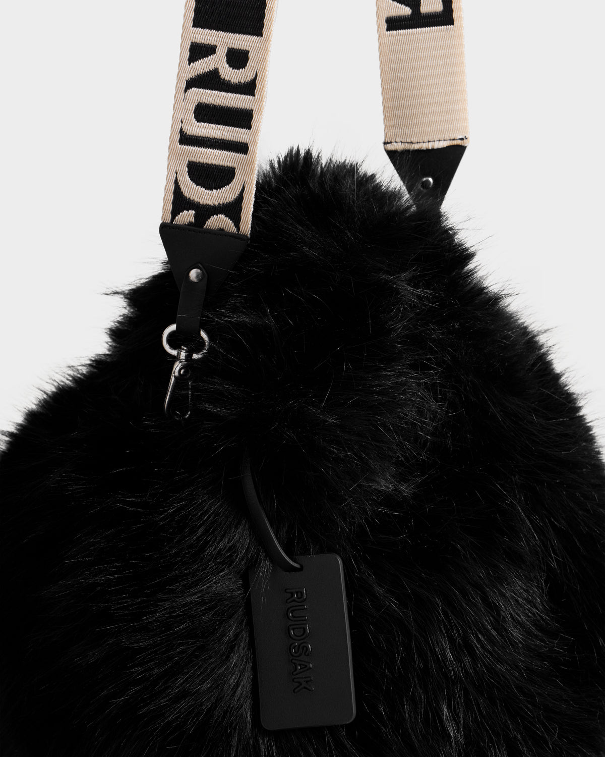 The CABAS - 8325545 Black by Rudsak is a black faux fur mini tote with a beige strap featuring “RUDSAK” in bold. A black leather Rudsak tag completes this statement accessory, ideal for elevating your winter look.