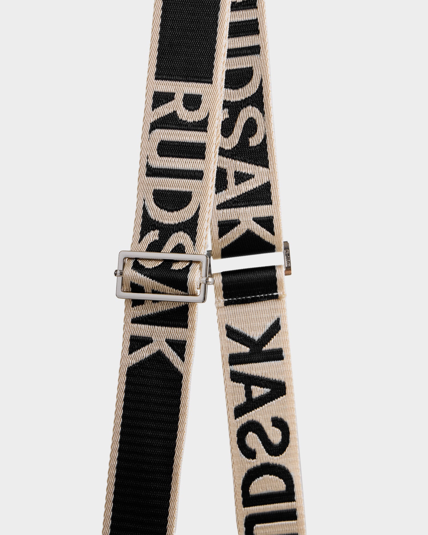 A close-up of CABAS - 8325545 Black by Rudsak shows a beige and black woven strap with "RUDSAK" in bold letters, featuring a silver rectangular buckle, set against a plain white background.