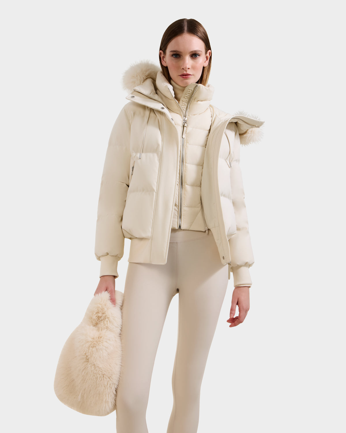 A woman in a cream puffer jacket, matching leggings, and holding the Rudsak CABAS - 8325545 Vanilla faux fur mini tote poses against a white background, highlighting chic winter style.