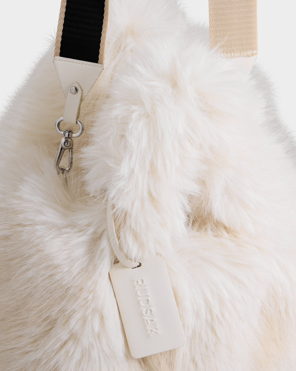A close-up of the CABAS - 8325545 Vanilla, a fluffy, cream-colored faux fur mini tote by Rudsak, featuring a beige brand tag, metal clasp, and black and beige strap—the perfect statement accessory for winter.