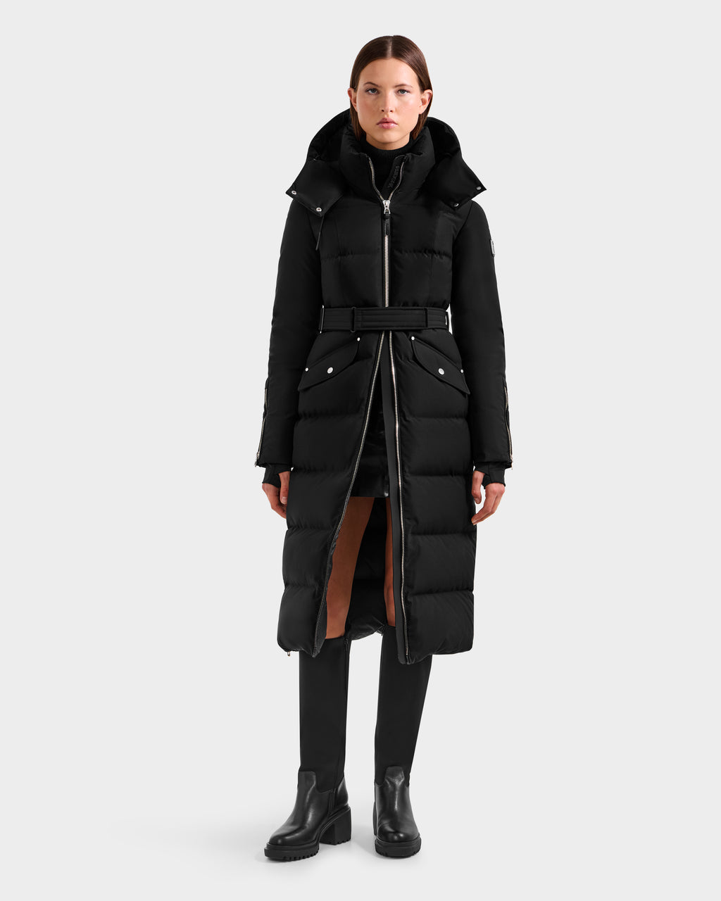 A woman faces forward in a Rudsak CALI H - 8125614 Black long down parka with high-loft down, belted waist, and high collar, paired with black pants and ankle boots against a plain light background.