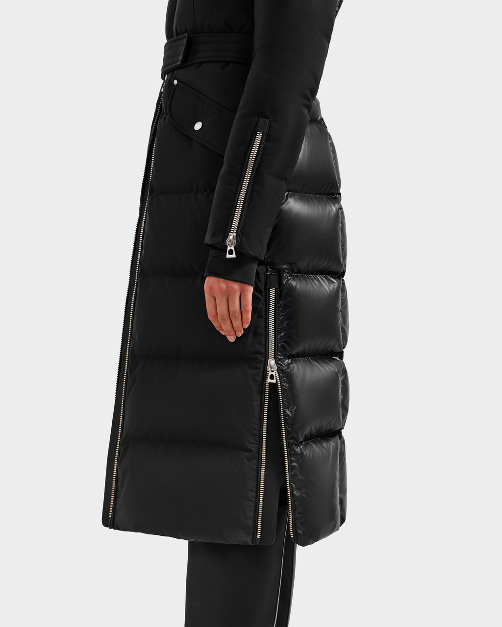 A person displays the lower half of the Rudsak CALI H - 8125614 Black women’s down parka, featuring visible side zippers, silver snap buttons, and high-loft down for warmth, with one hand shown against a plain background.
