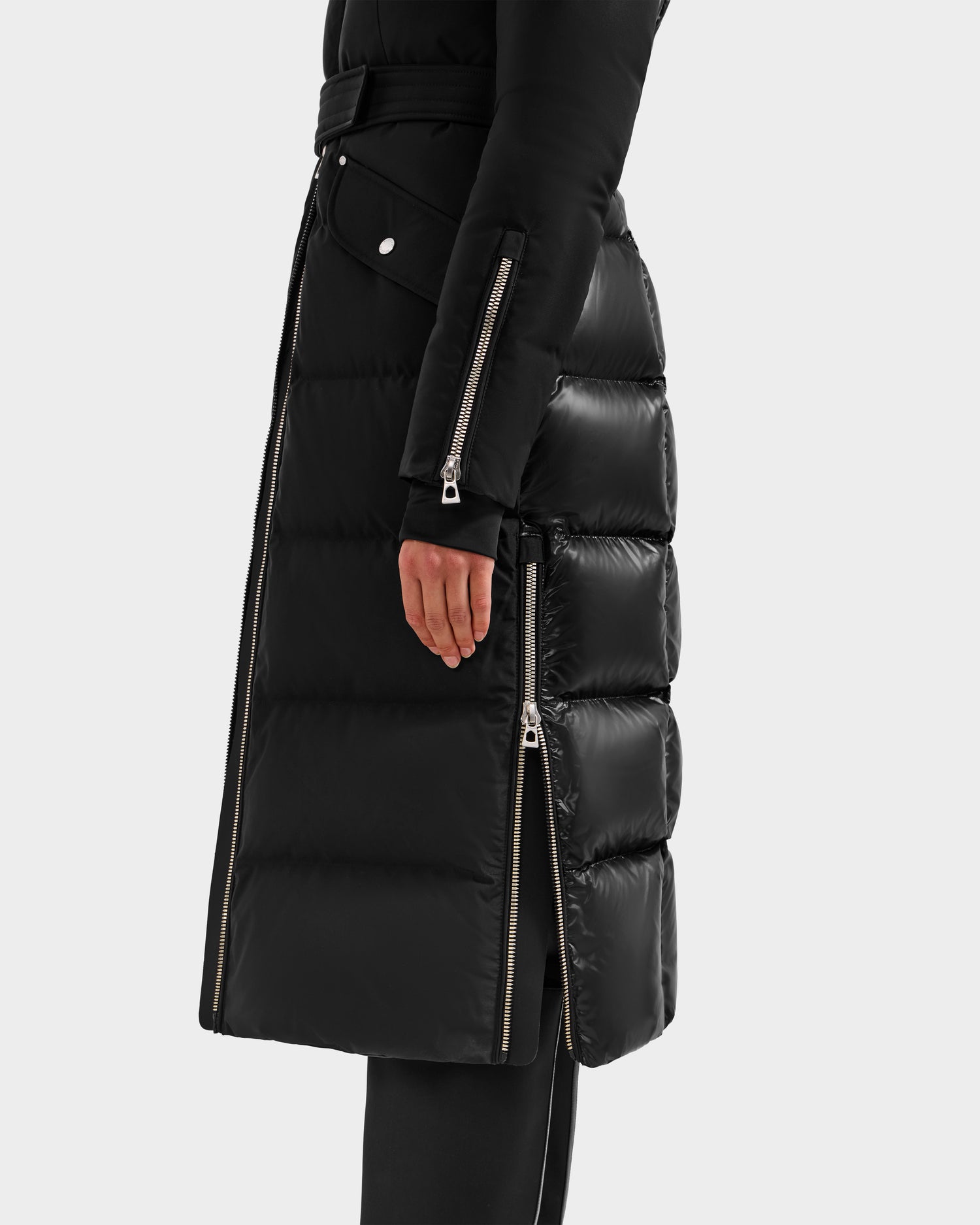 A person displays the lower half of the Rudsak CALI H - 8125614 Black women’s down parka, featuring visible side zippers, silver snap buttons, and high-loft down for warmth, with one hand shown against a plain background.