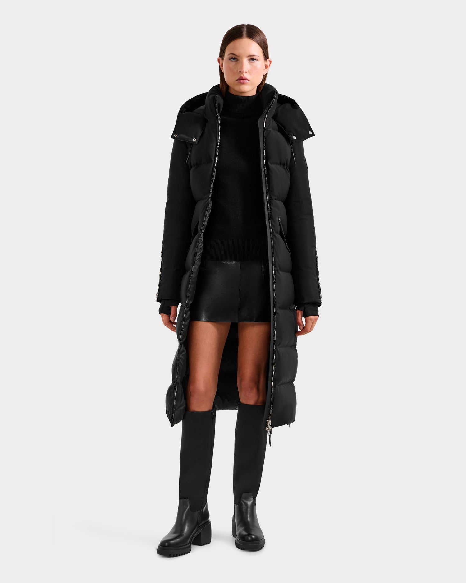 A woman faces forward, wearing the Rudsak CALI H - 8125614 Black women’s long down parka with a removable hood, styled over a black turtleneck, mini skirt, and knee-high boots against a light gray background.