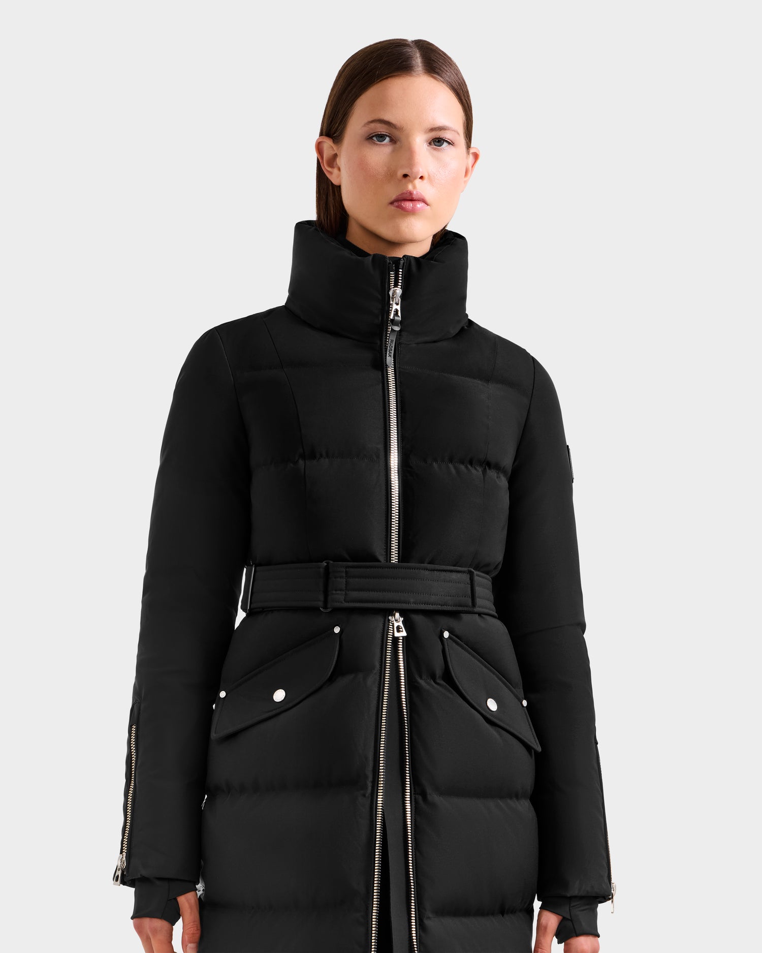 A woman wearing the Rudsak CALI H - 8125614 Black belted long down parka with large front pockets, high collar, and high-loft down stands against a plain light background.