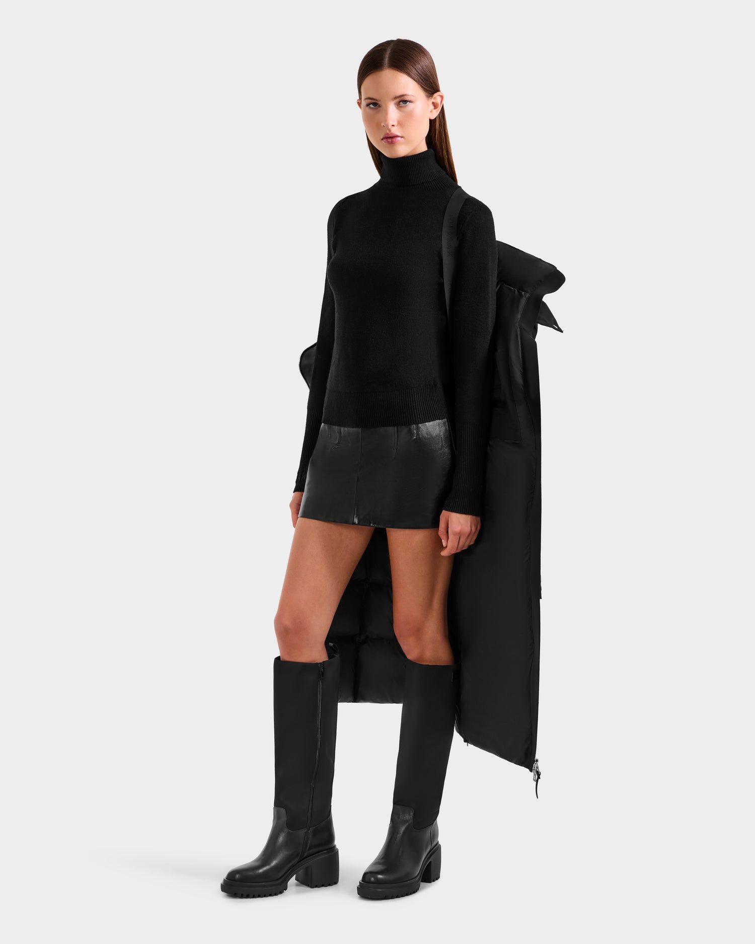 A woman stands against a plain background wearing the Rudsak CALI H - 8125614 Black women’s long down parka with removable hood draped over her shoulders, styled with a black turtleneck, leather mini skirt, and knee-high black boots.