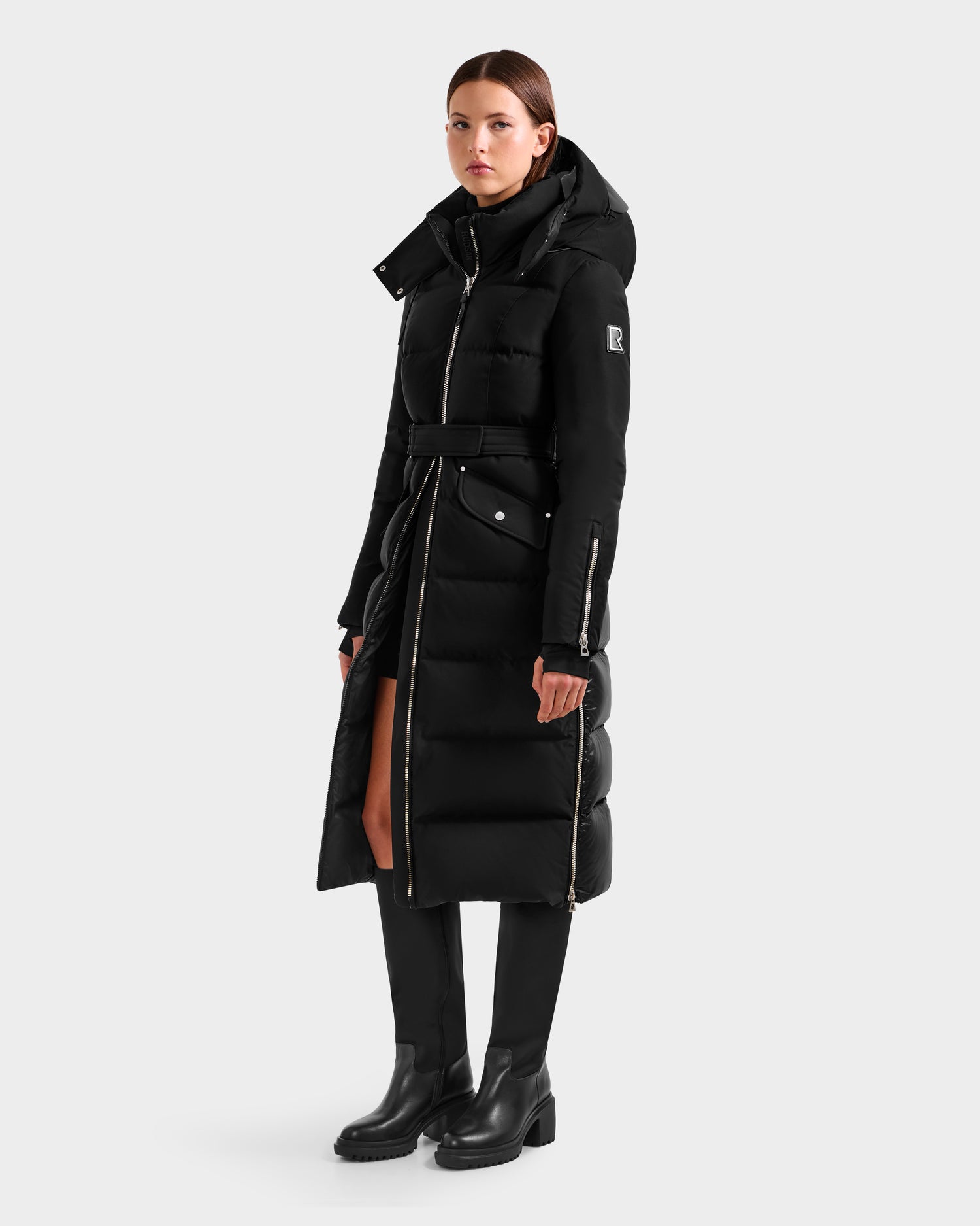 A woman wears the Rudsak CALI H - 8125614 Black women's long down parka with a removable hood, belt, and zipper details, paired with knee-high black boots, set against a plain white background.