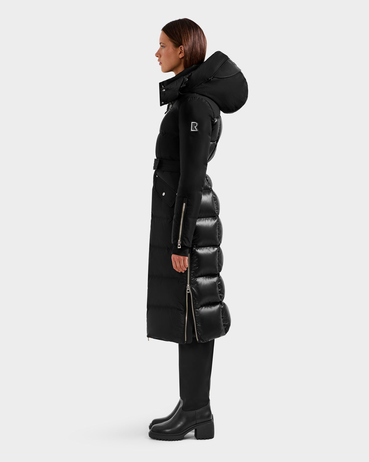 A woman in profile wears the Rudsak CALI H - 8125614 Black women’s long down parka with removable hood, waist belt, and side zippers, paired with black heeled boots, set against a plain light background.