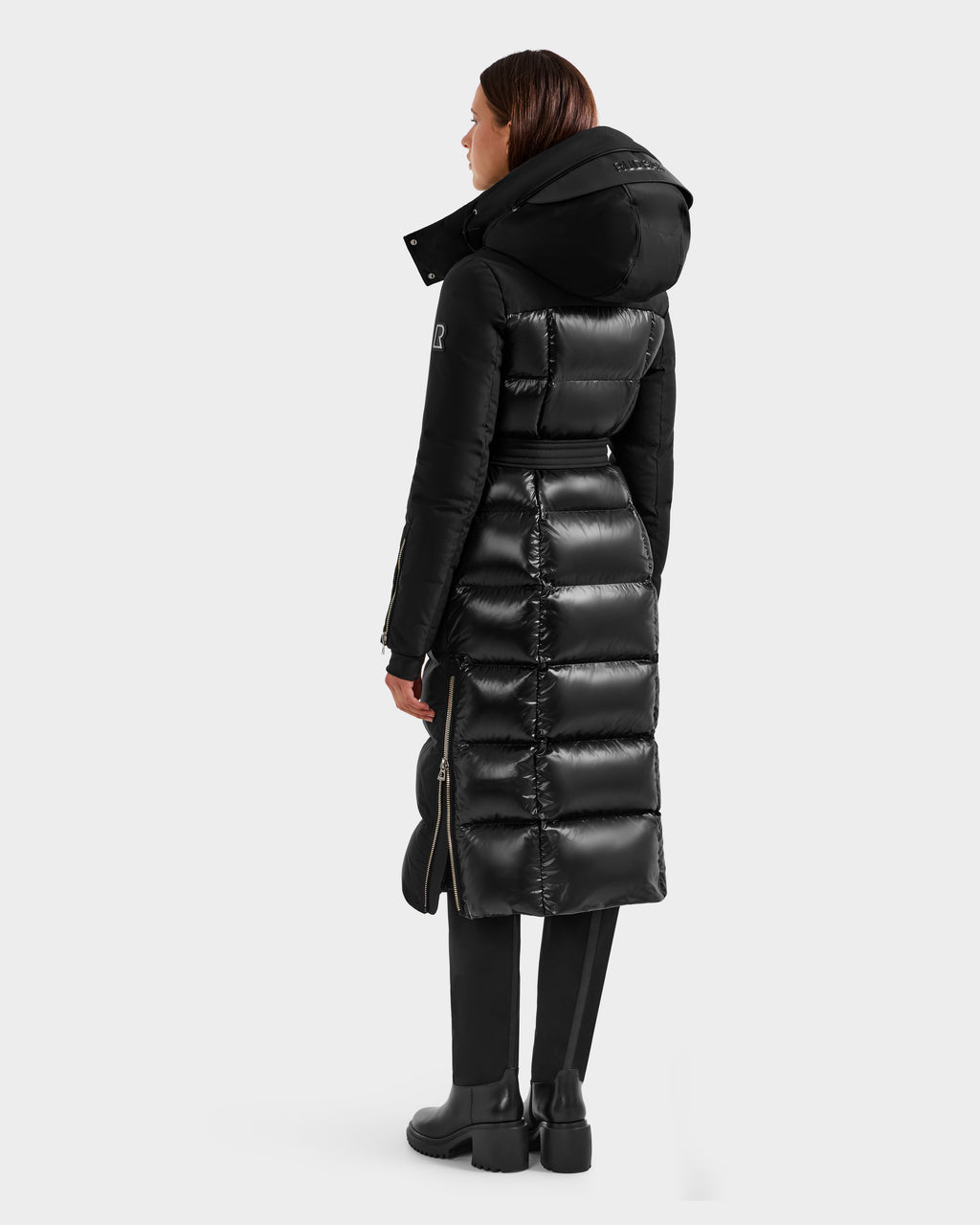 A woman faces away, wearing the Rudsak CALI H - 8125614 Black long down parka with high-loft fill and side zippers, paired with black pants and boots, against a plain light background.