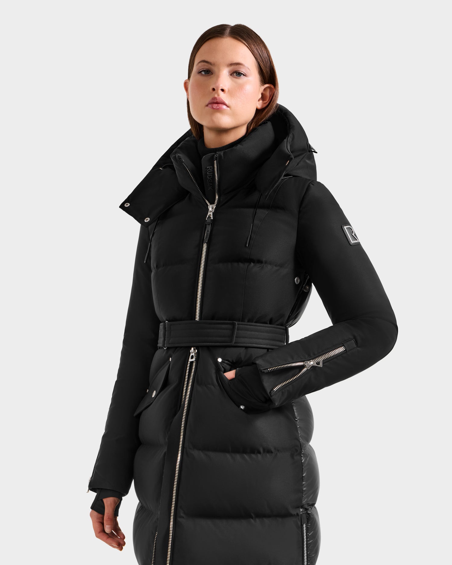 A woman stands facing forward with one hand in her pocket, wearing the Rudsak CALI H - 8125614 Black women’s long down parka with a high collar, belt, and removable hood against a plain light background.
