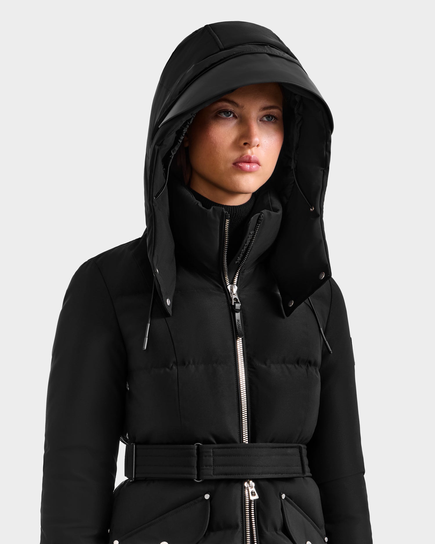 A person stands against a plain light background wearing the Rudsak CALI H - 8125614 Black women’s long down parka, featuring a removable hood, silver buttons, drawstrings, zipper, and belt for added style and warmth.