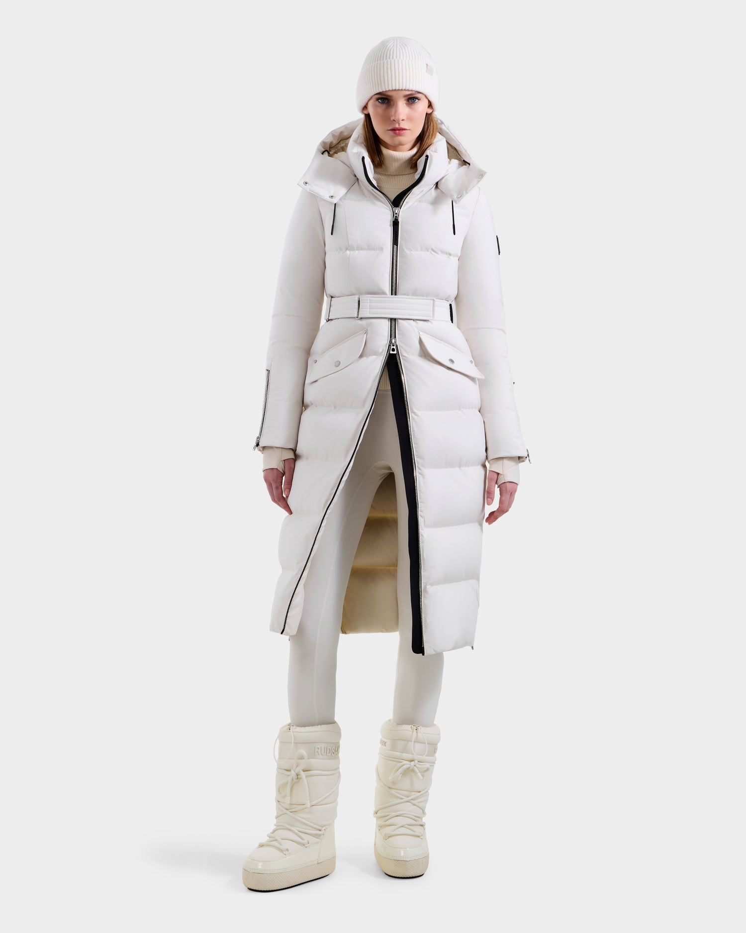 A woman faces forward wearing the Rudsak CALI H - 8125614 Vanilla women's long down parka with high-loft 800+ fill power down and removable hood, paired with a white knit beanie, white leggings, and snow boots against a light background.