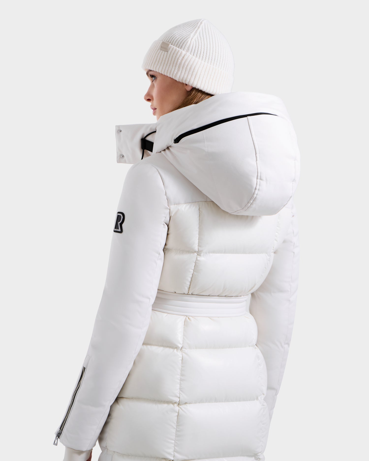 A woman faces away against a light gray background, wearing the Rudsak CALI H - 8125614 Vanilla women's long white down parka with a removable hood and 800+ fill power, paired with a white knit beanie.