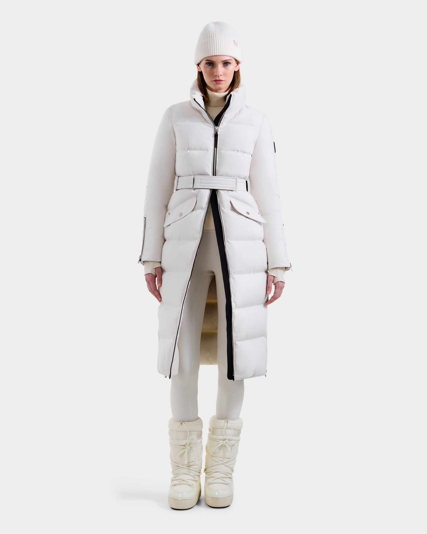A person stands facing forward, wearing the Rudsak CALI H - 8125614 Vanilla women’s long down parka, belted at the waist, paired with white leggings, winter boots, and a knit beanie against a plain white background.