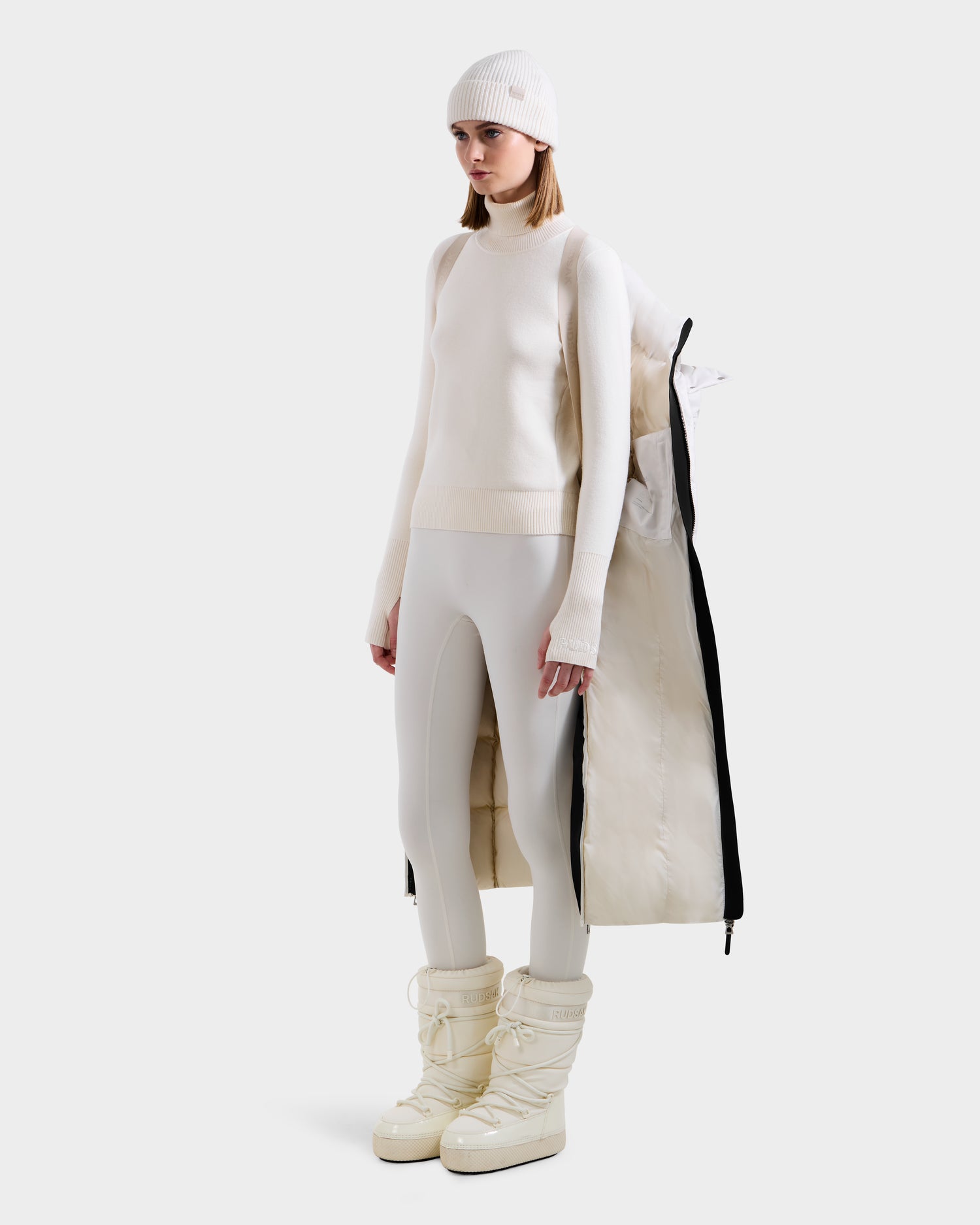 A woman wears the Rudsak CALI H - 8125614 Vanilla women’s long down parka with removable hood, styled over cream knitwear and off-white boots, standing against a white background.