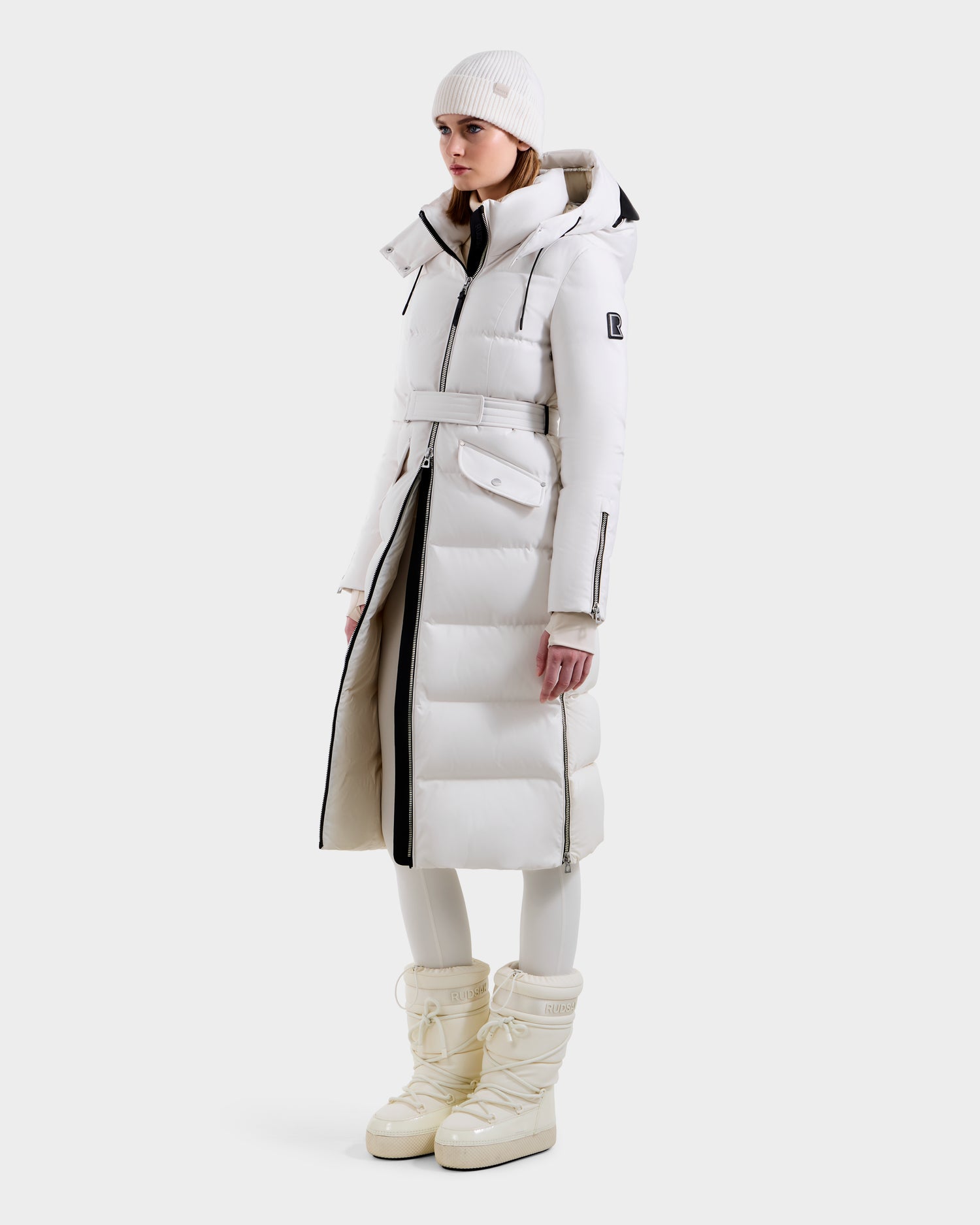 A woman stands against a plain background wearing the Rudsak CALI H - 8125614 Vanilla women’s long down parka with a belt and removable hood, paired with white knit beanie, white leggings, and cream snow boots, looking forward with hands by her sides.