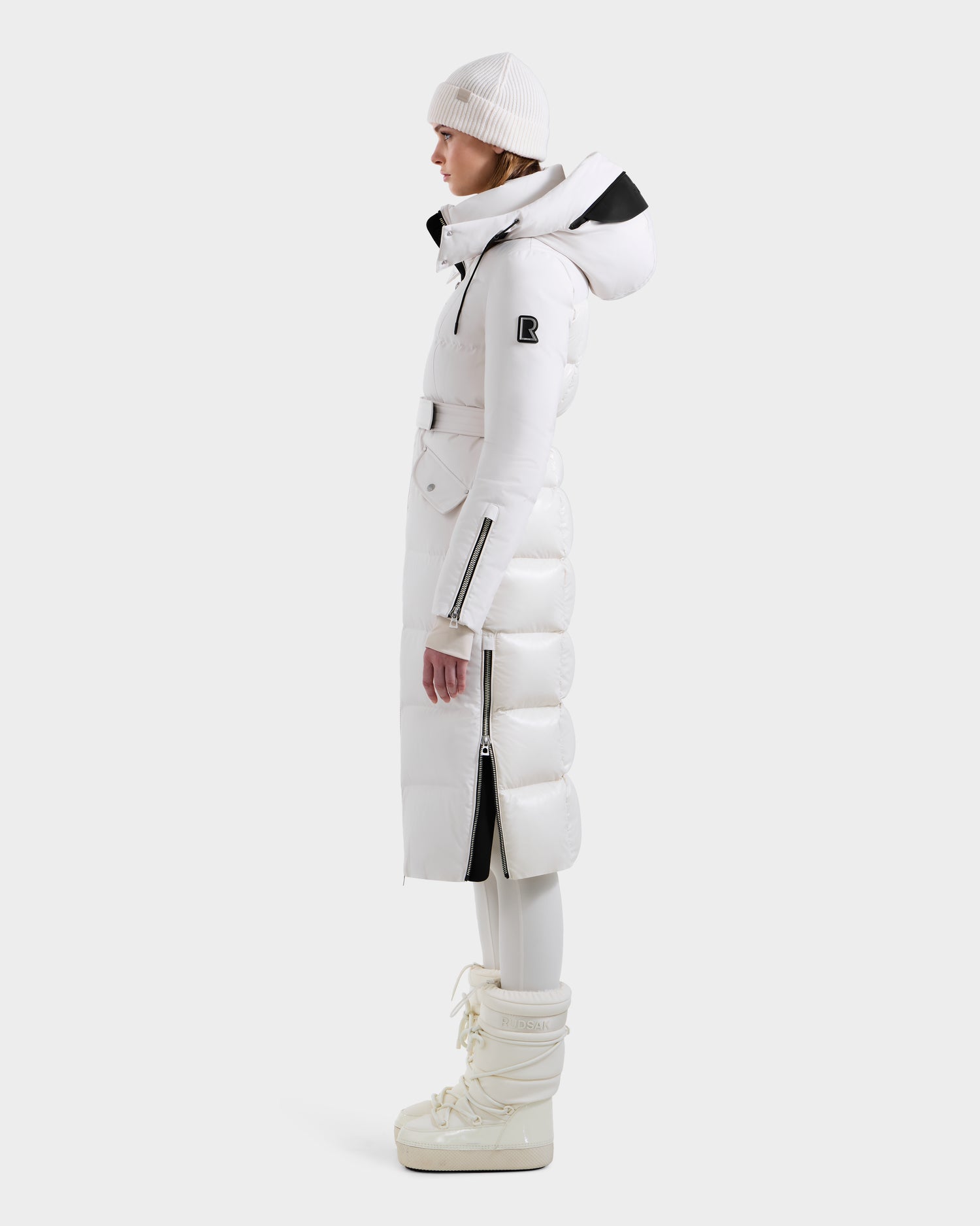 A person in profile wears the Rudsak CALI H - 8125614 Vanilla women’s long down parka with a hood, paired with a white beanie, pants, and winter boots, standing against a plain light backdrop.