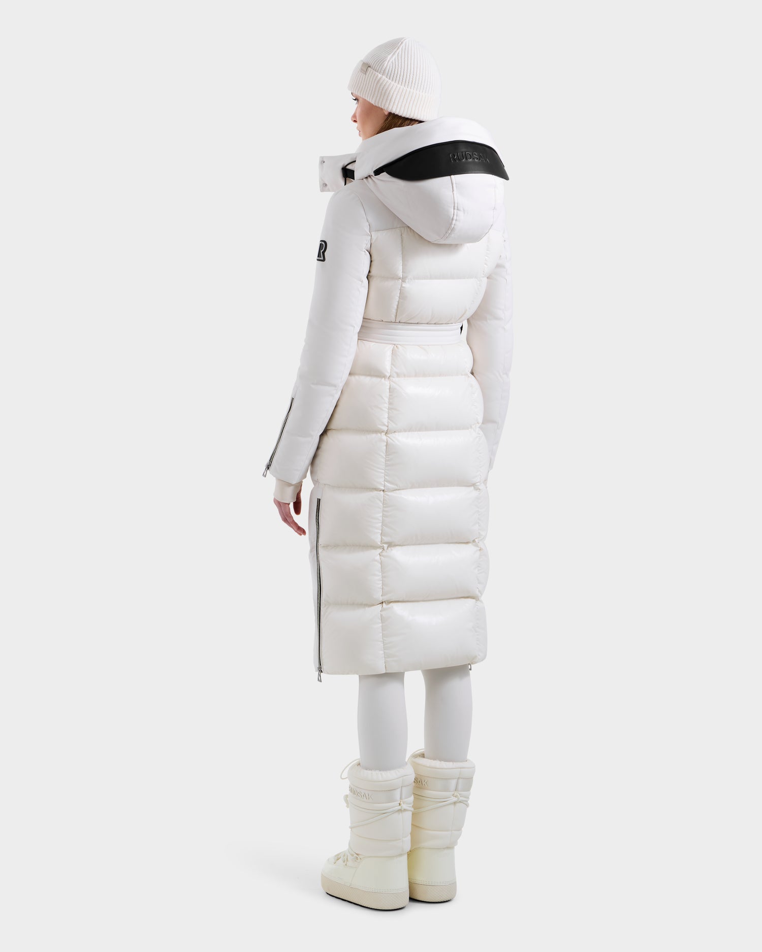 A person in the Rudsak CALI H - 8125614 Vanilla women’s long down parka with 800+ fill power, black-trimmed removable hood, white pants, boots, and beanie stands facing away against a plain, light background.
