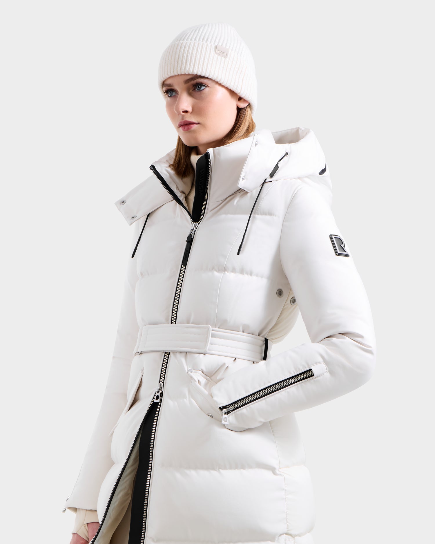 A woman wears the Rudsak CALI H - 8125614 Vanilla women’s long down parka in white, featuring a belted waist, removable hood, black zippers, 800+ fill power down, and sleeve logo patch. Styled with a knit beanie on a light gray background.