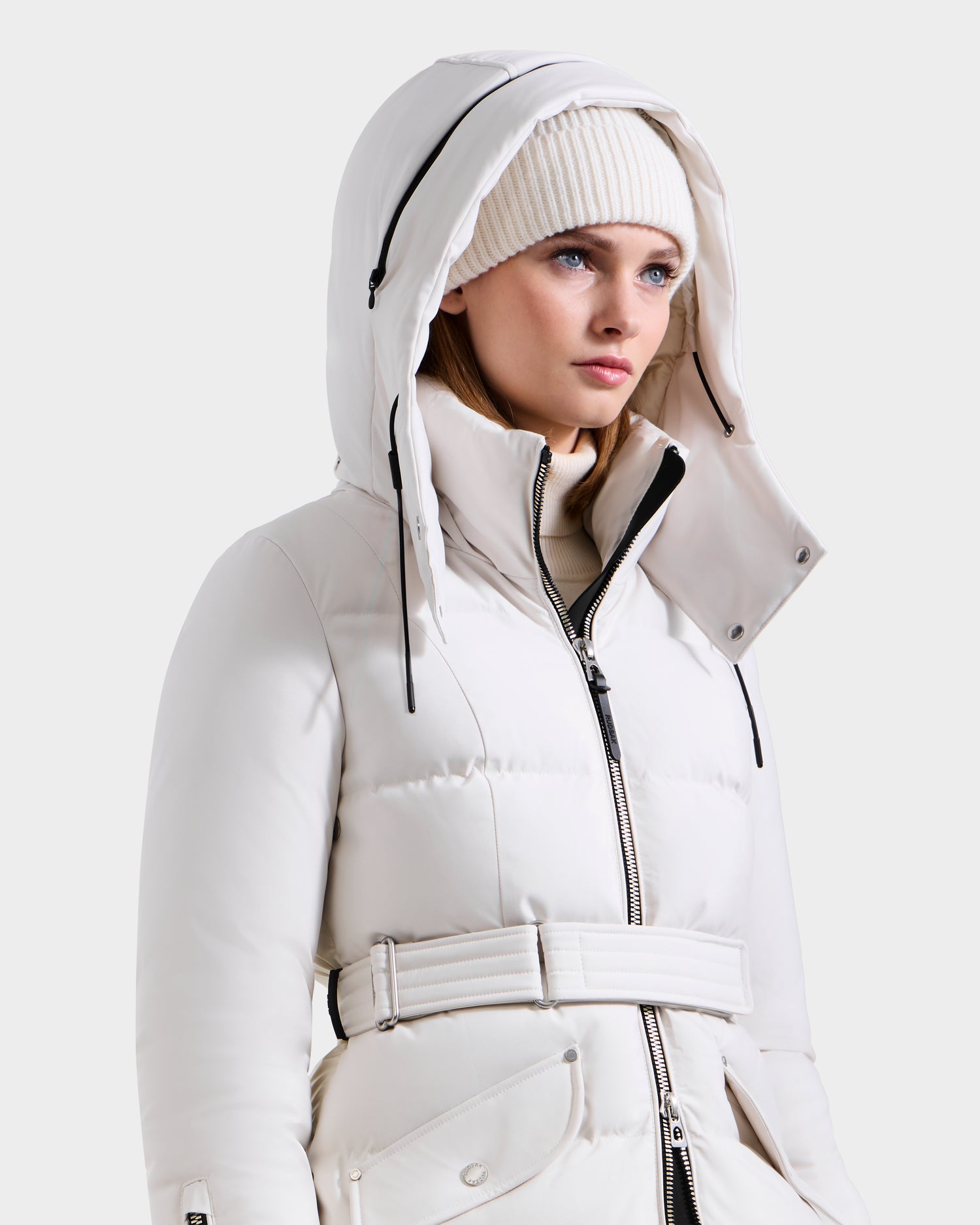 A woman wears the Rudsak CALI H - 8125614 Vanilla belted long down parka with high-loft 800+ fill power down and a removable hood, paired with a white knit beanie, looking slightly to the side against a plain light background.