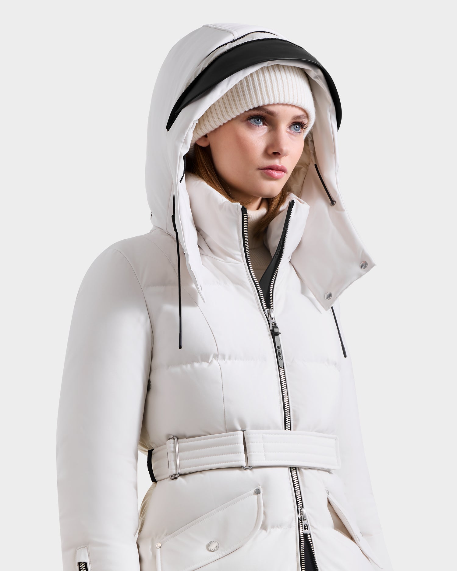 A woman models the Rudsak CALI H - 8125614 Vanilla women’s long down parka with belt and removable hood up, paired with a white knit beanie, gazing slightly to the side against a plain light background.