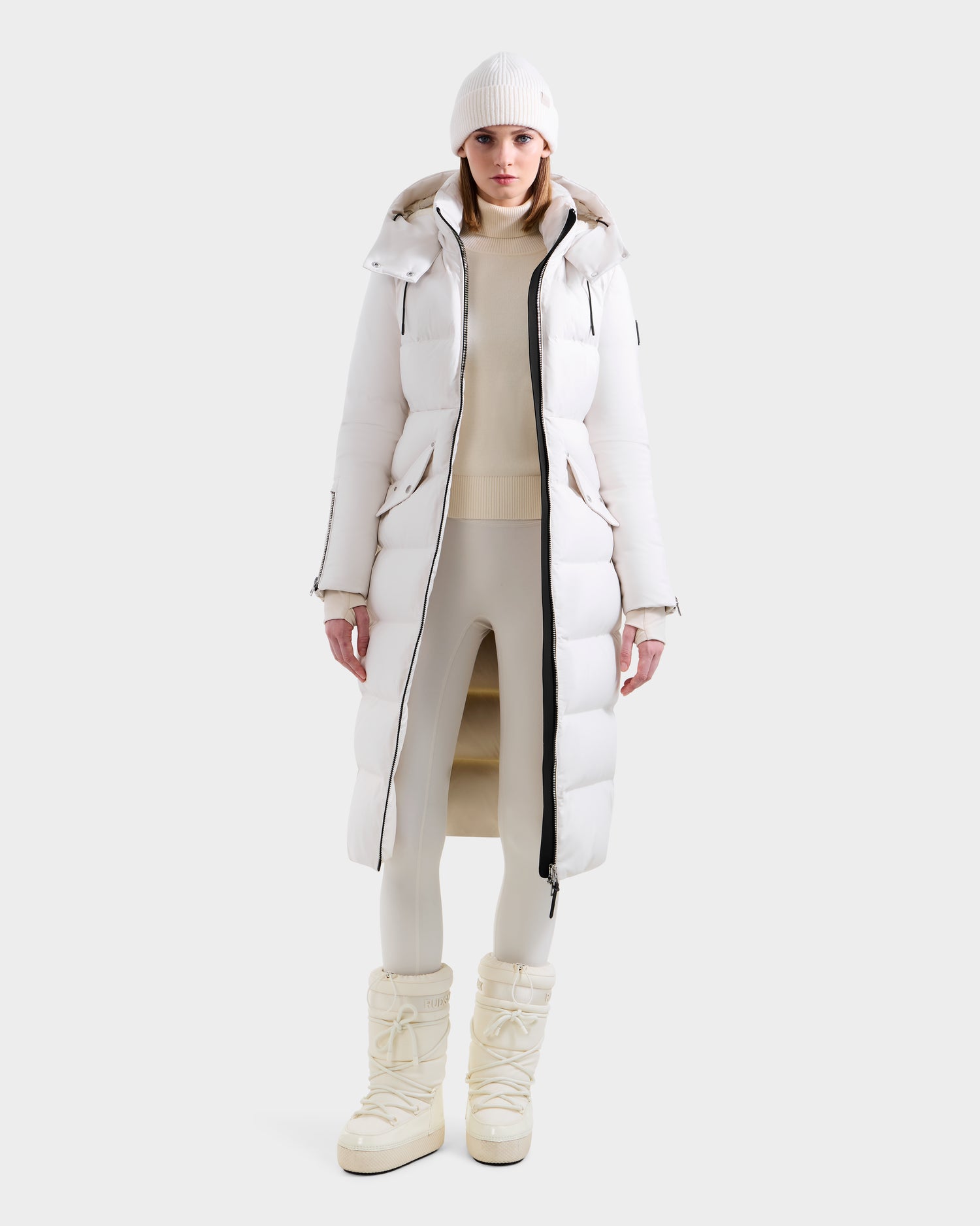 A person wearing the Rudsak CALI H - 8125614 Vanilla women’s long down parka stands facing forward in a light background, styled with a light beige turtleneck, matching leggings, white beanie, and off-white winter boots.