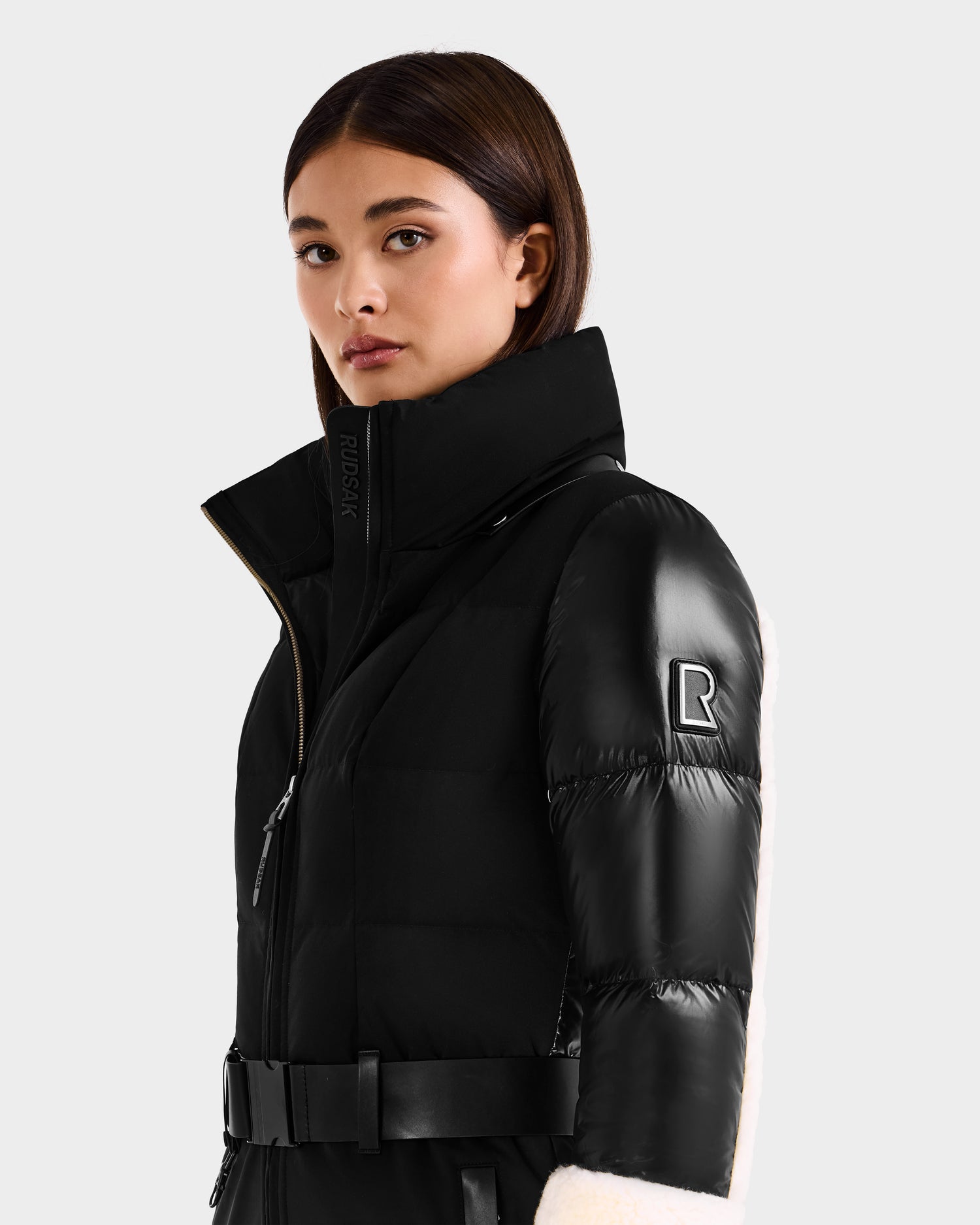 A woman wears the Rudsak CARMEN - 8125708 puffer jacket in black, featuring a belt, high collar, and R logo patch on the sleeve, as she stands confidently against a plain light gray background.