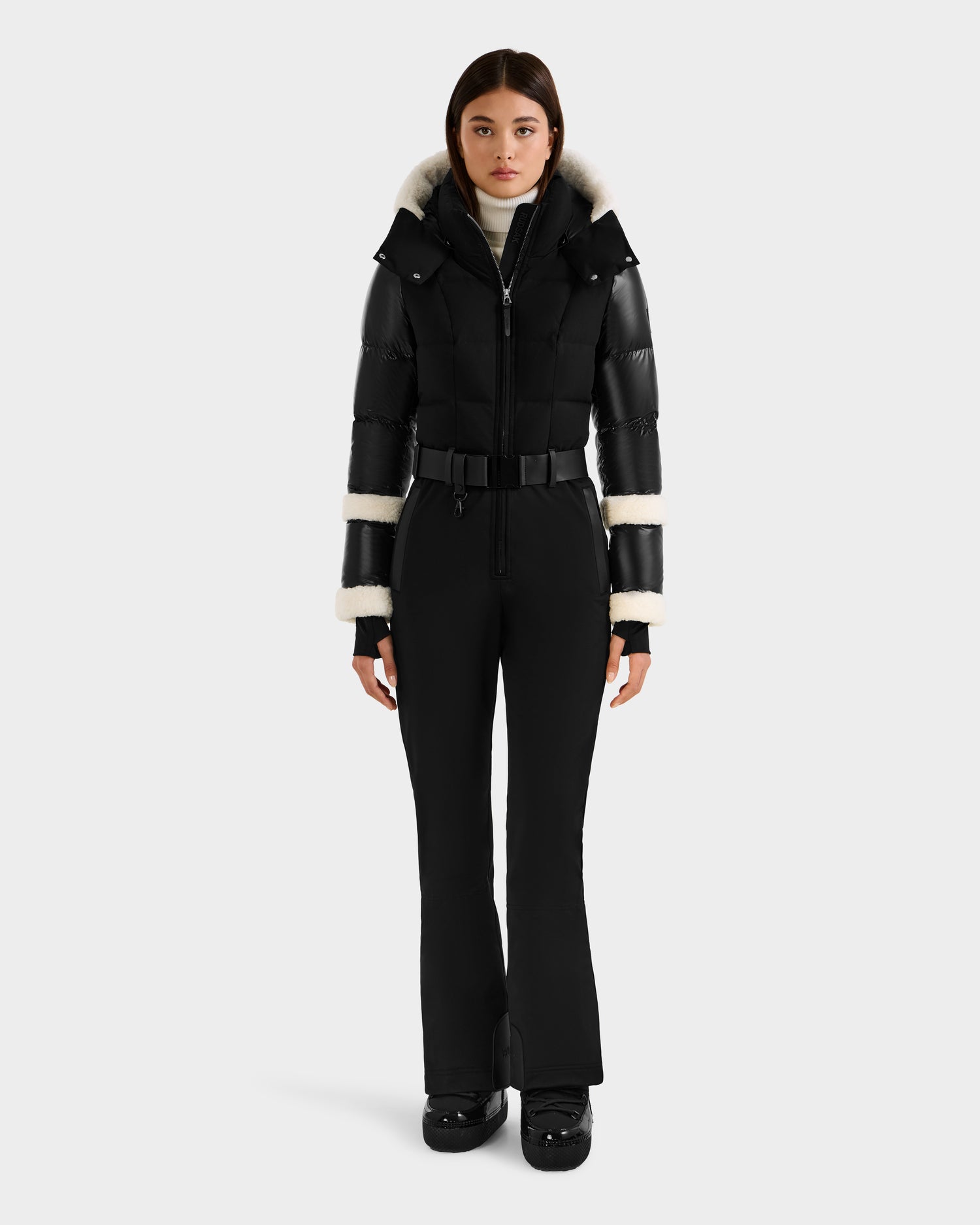 A woman stands facing forward, wearing the Rudsak CARMEN - 8125708 Black one-piece ski suit with white fur trim on the hood and sleeves, paired with black gloves and boots, against a plain white background.