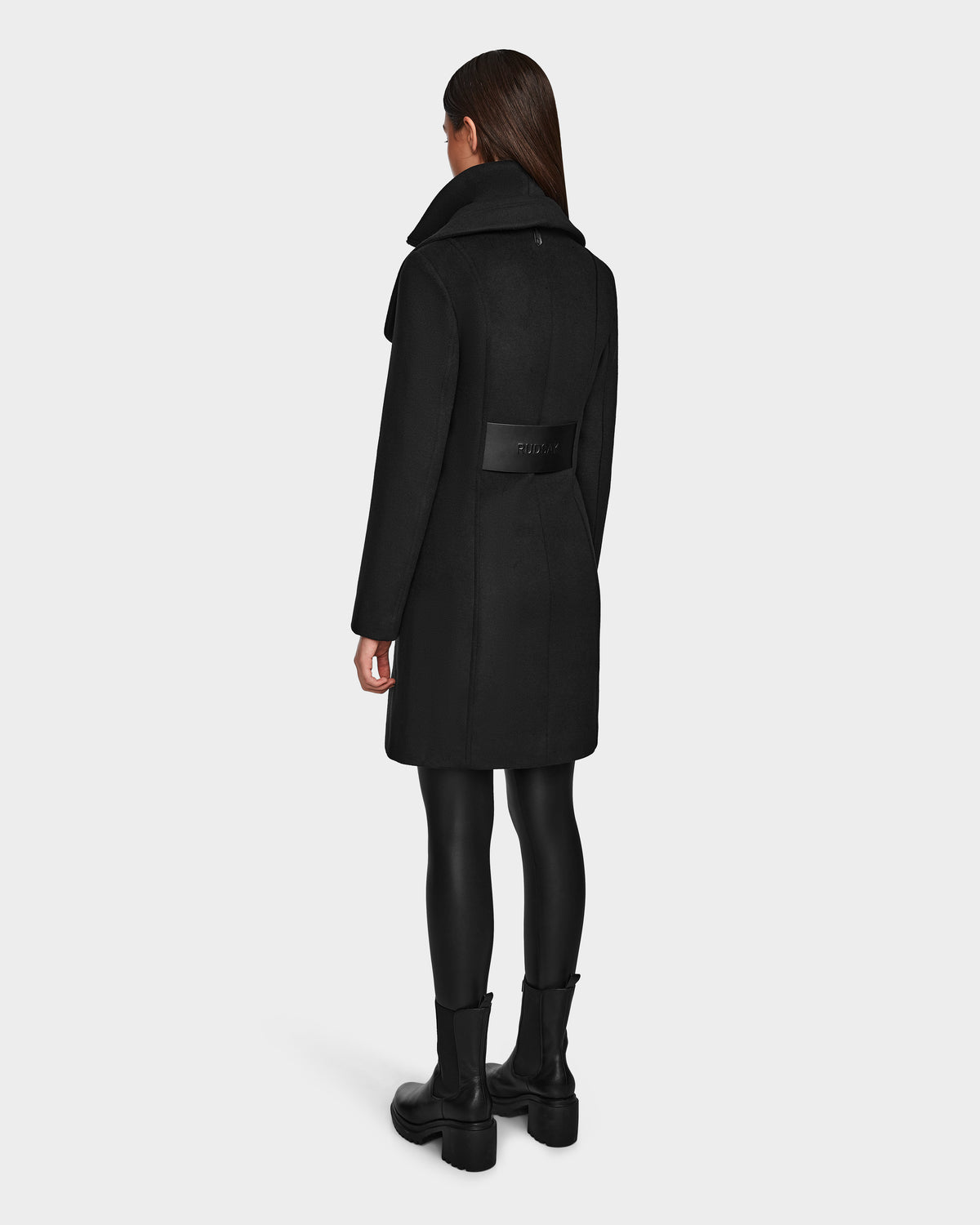 A woman with long brown hair, wearing the Rudsak CAROLINE - L124543 Black premium wool blend coat, black leggings, and black boots, stands facing away from the camera against a plain light gray background.