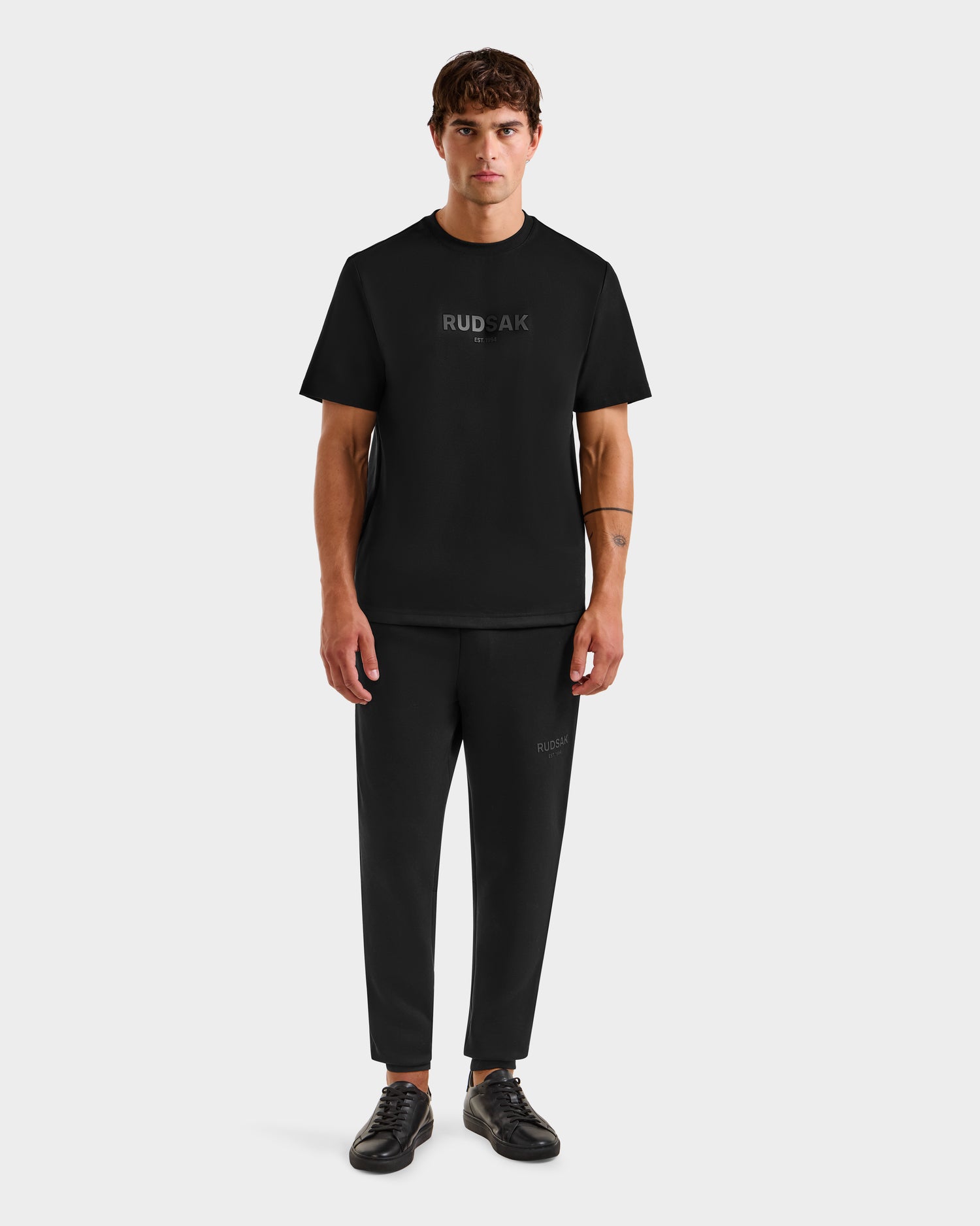 A man faces forward, wearing the Rudsak CASEY - 6525037 Black T-shirt and black pants with Rudsak logos, paired with black sneakers, set against a plain white background.