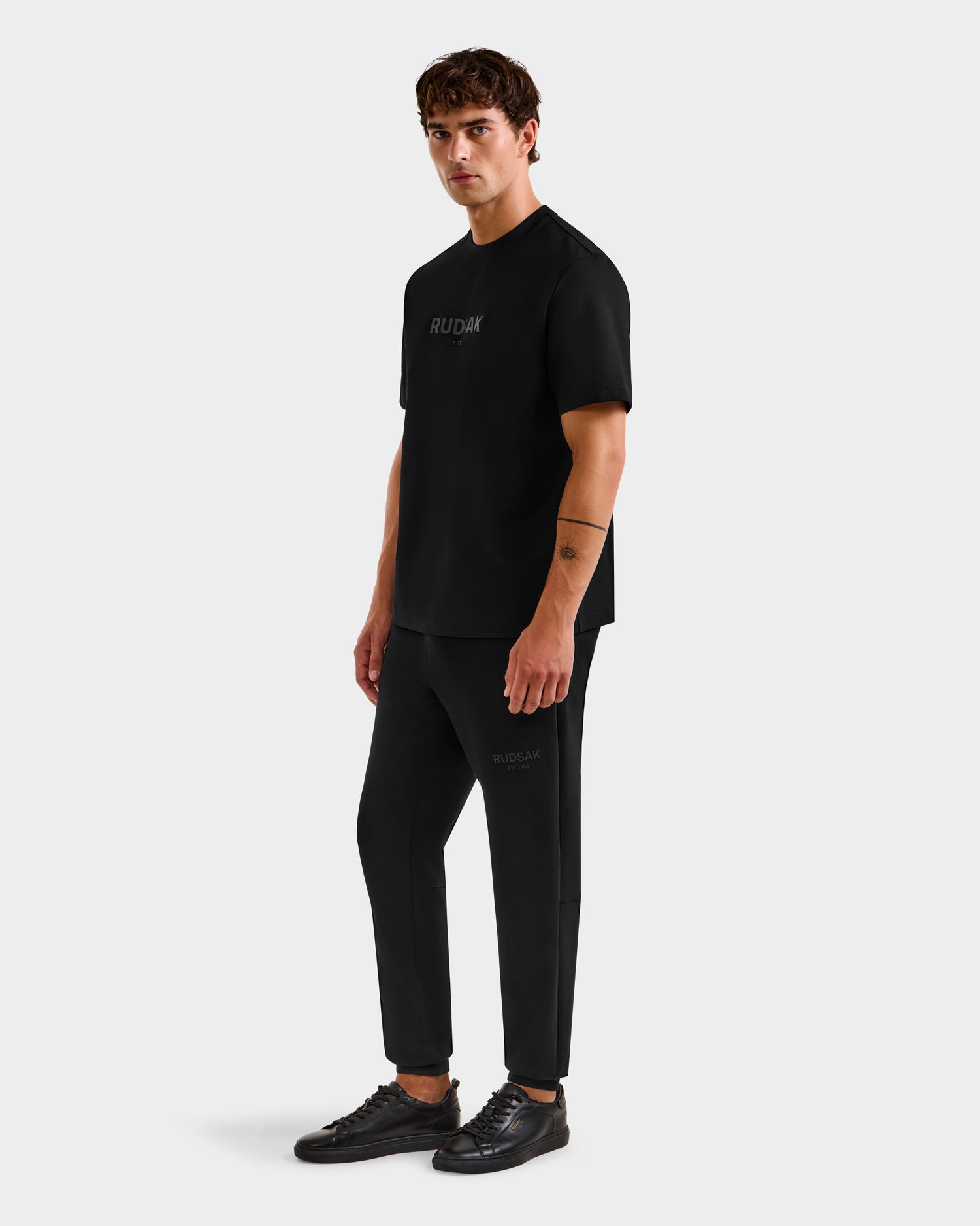 A man stands facing slightly to the side, wearing the Rudsak CASEY - 6525037 Black mens T-shirt, with matching black pants and sneakers, against a plain light gray background.