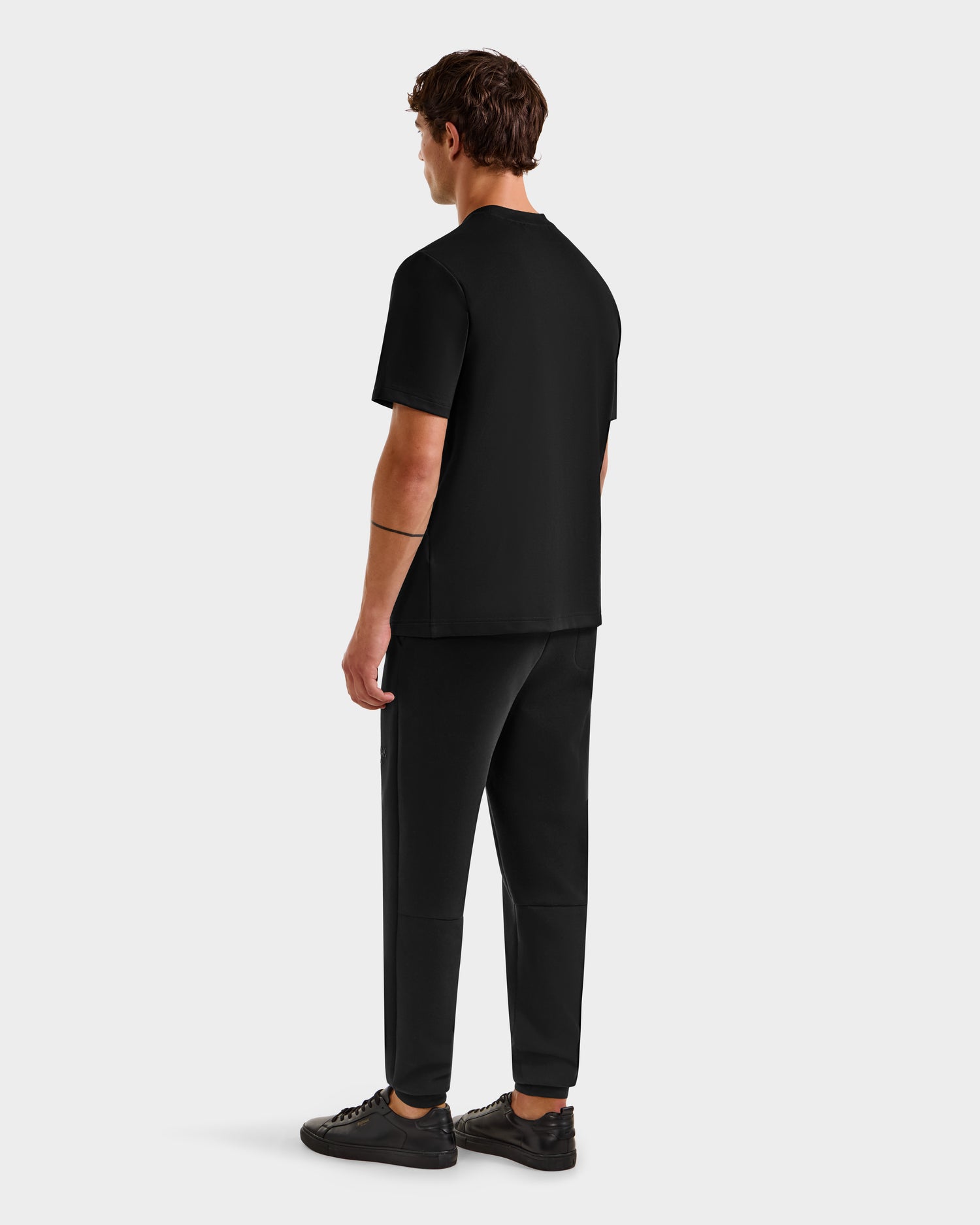 A man stands facing away, wearing the Rudsak CASEY - 6525037 Black T-shirt, black pants, and black sneakers against a plain light gray background.