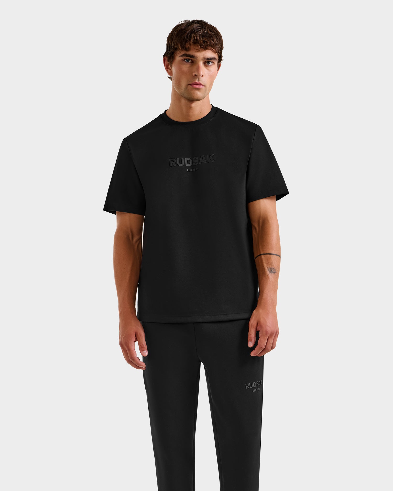 A man with short brown hair wears the Rudsak CASEY - 6525037 Black T-shirt and black pants with subtle branding, standing against a plain light gray background and facing forward with a neutral expression for a comfortable, essential look.