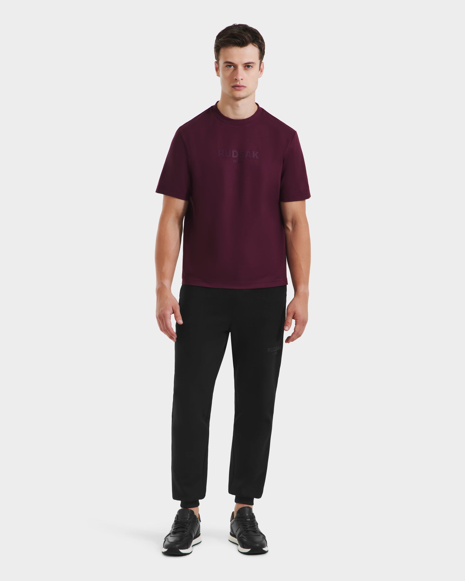 A man stands facing forward, wearing the Rudsak CASEY - 6525037 Bordeaux short-sleeve T-shirt with black pants and black sneakers against a plain white background.