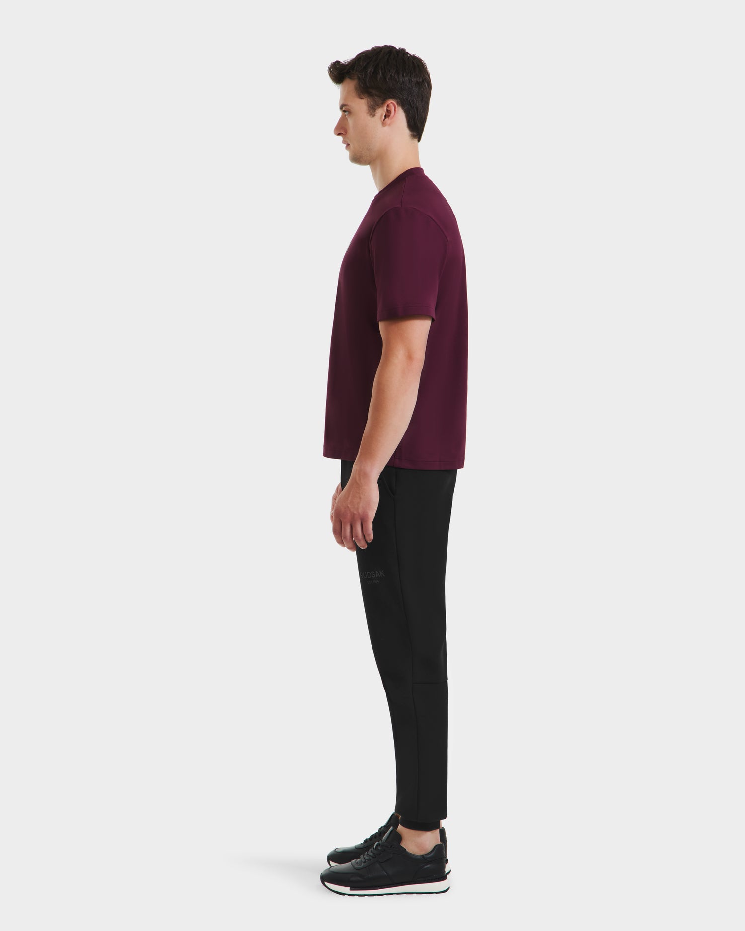 A man stands in profile facing left, wearing the Rudsak CASEY - 6525037 Bordeaux T-shirt—an undeniably comfortable essential—paired with black pants and black sneakers against a plain white background.
