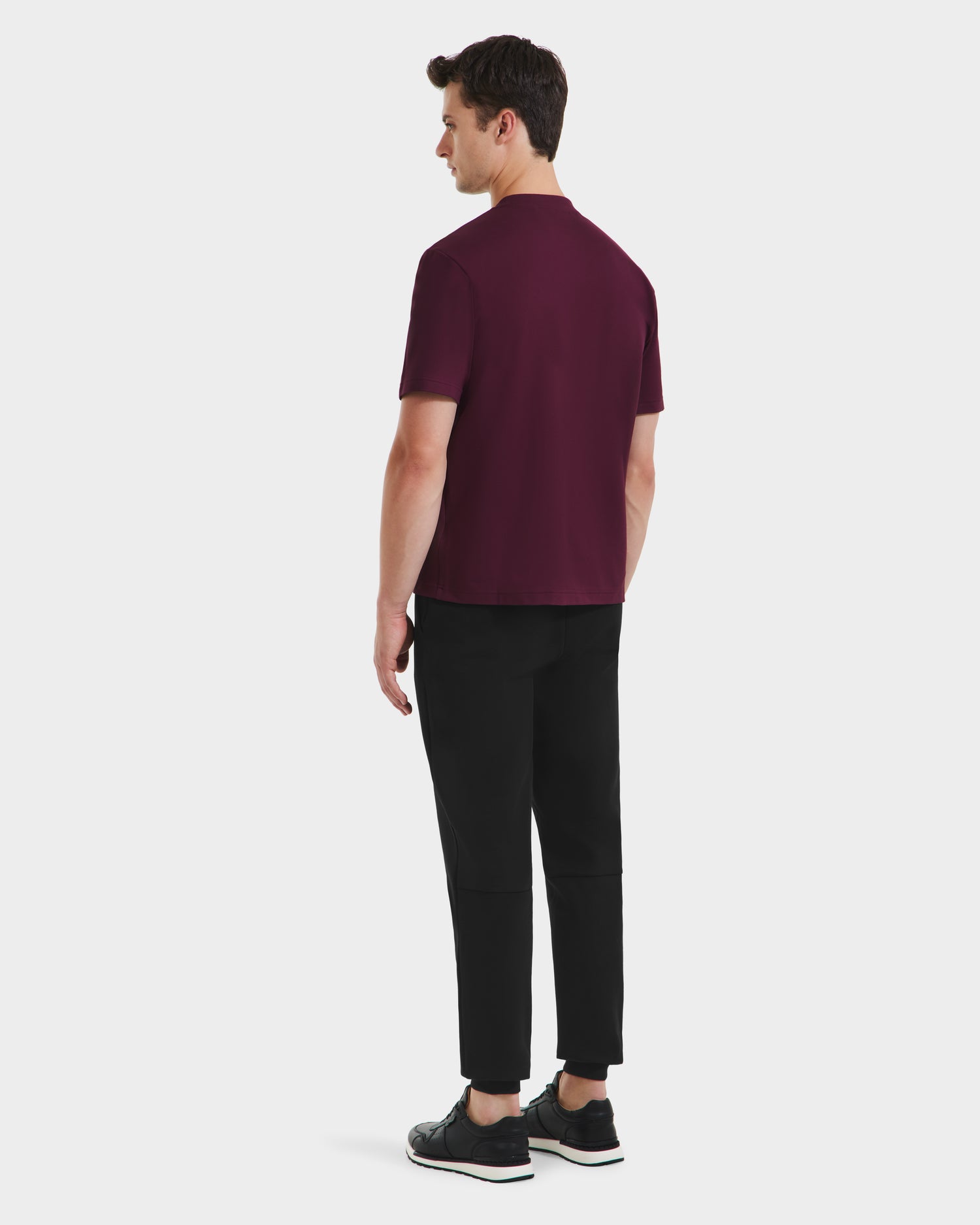 A man faces away, wearing the Rudsak CASEY - 6525037 Bordeaux T-shirt with black pants and shoes, showcasing this comfortable essential against a plain white background.