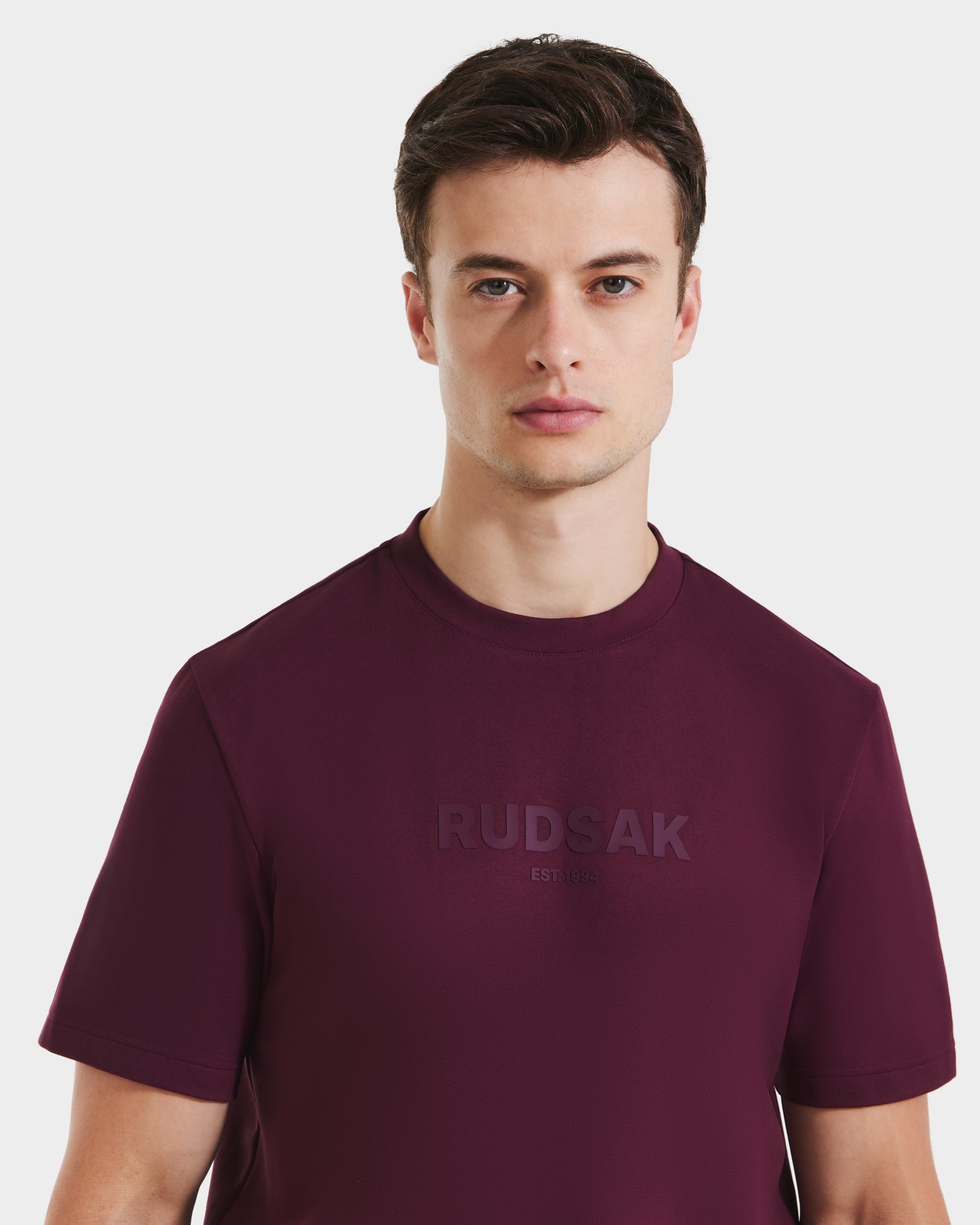 A young man with short brown hair wears the Rudsak CASEY - 6525037 Bordeaux men's T-shirt, standing against a plain white background and looking directly at the camera with a neutral expression.