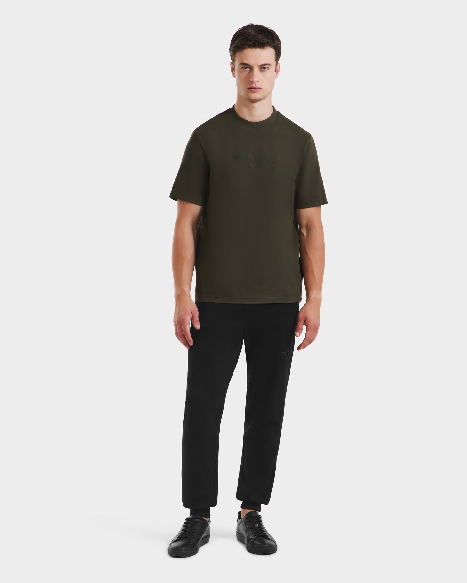 A young man faces forward, wearing the Rudsak CASEY - 6525037 T-shirt in dark olive, paired with black pants and sneakers against a plain white background.