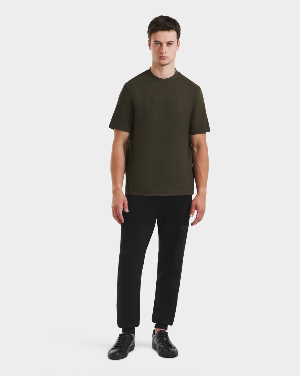 A young man faces forward, wearing the Rudsak CASEY - 6525037 T-shirt in dark olive, paired with black pants and sneakers against a plain white background.