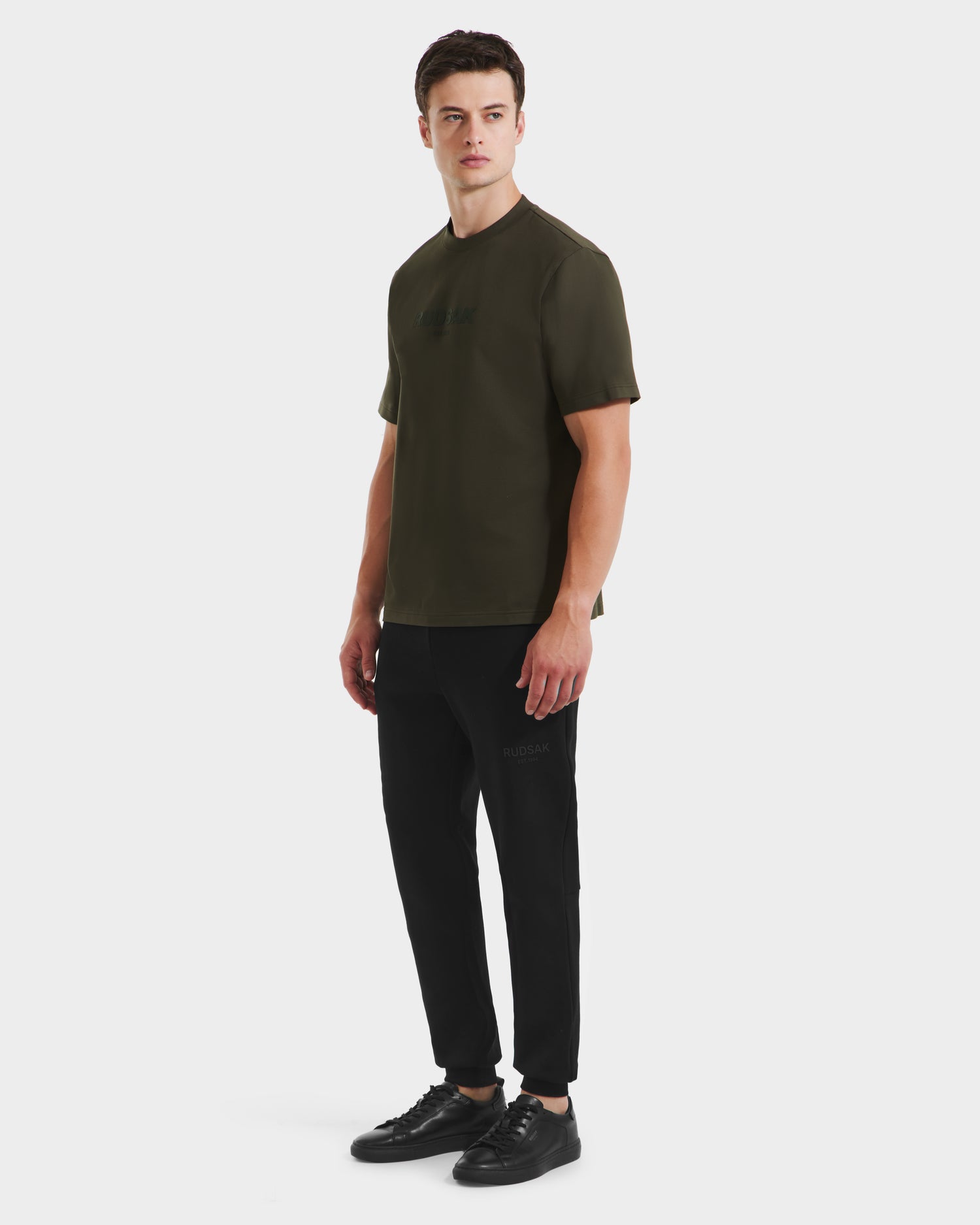 A young man stands against a white background wearing the Rudsak CASEY - 6525037 T-shirt in Dark Olive, with black jogger pants and black sneakers. He is facing slightly to the side with a neutral expression.