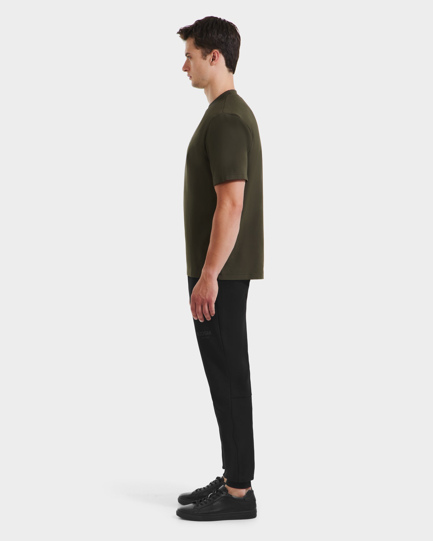 A man in profile facing left wears the Rudsak CASEY - 6525037 Dark Olive T-shirt, paired with black joggers and black sneakers, set against a plain white background.
