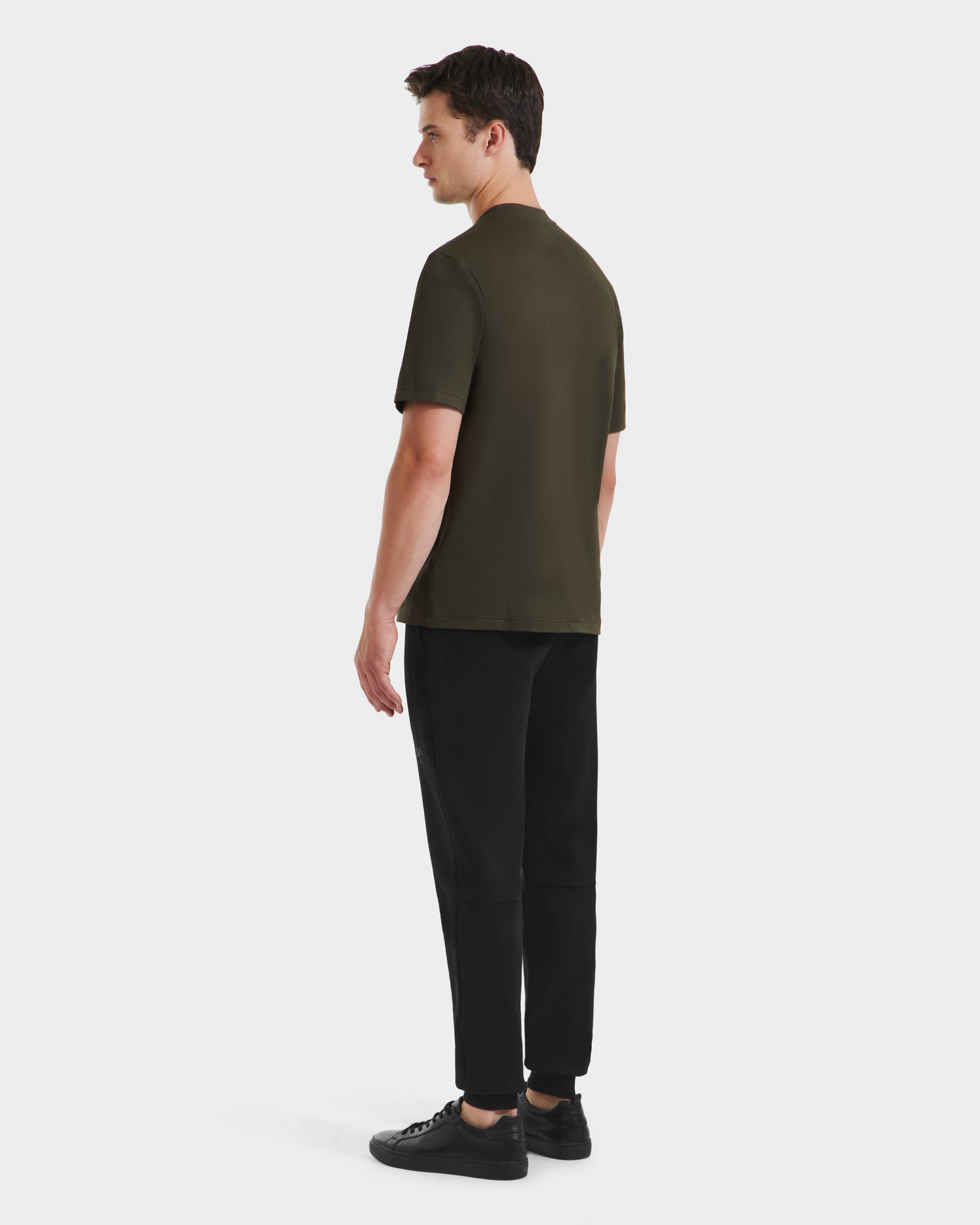 A man faces away, wearing the Rudsak CASEY - 6525037 Dark Olive mens T-shirt, paired with black pants and black sneakers, against a plain white background.