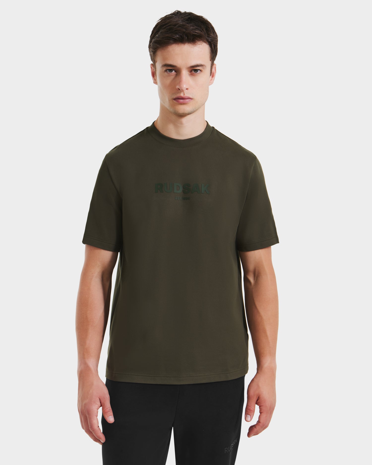 A young man stands facing forward in a dark olive Rudsak CASEY T-shirt with subtle chest branding, paired with black pants, against a plain white background.